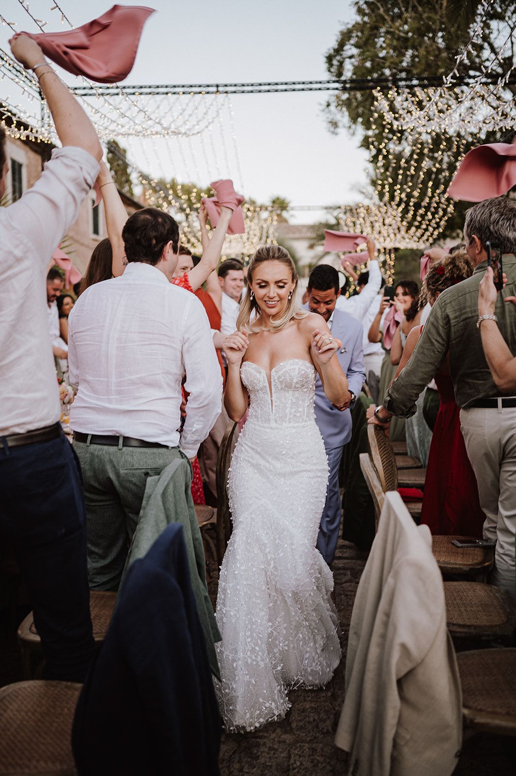 Bride in fitted sparkle gown walks joyfully through cheering guests under string lights at outdoor wedding reception.