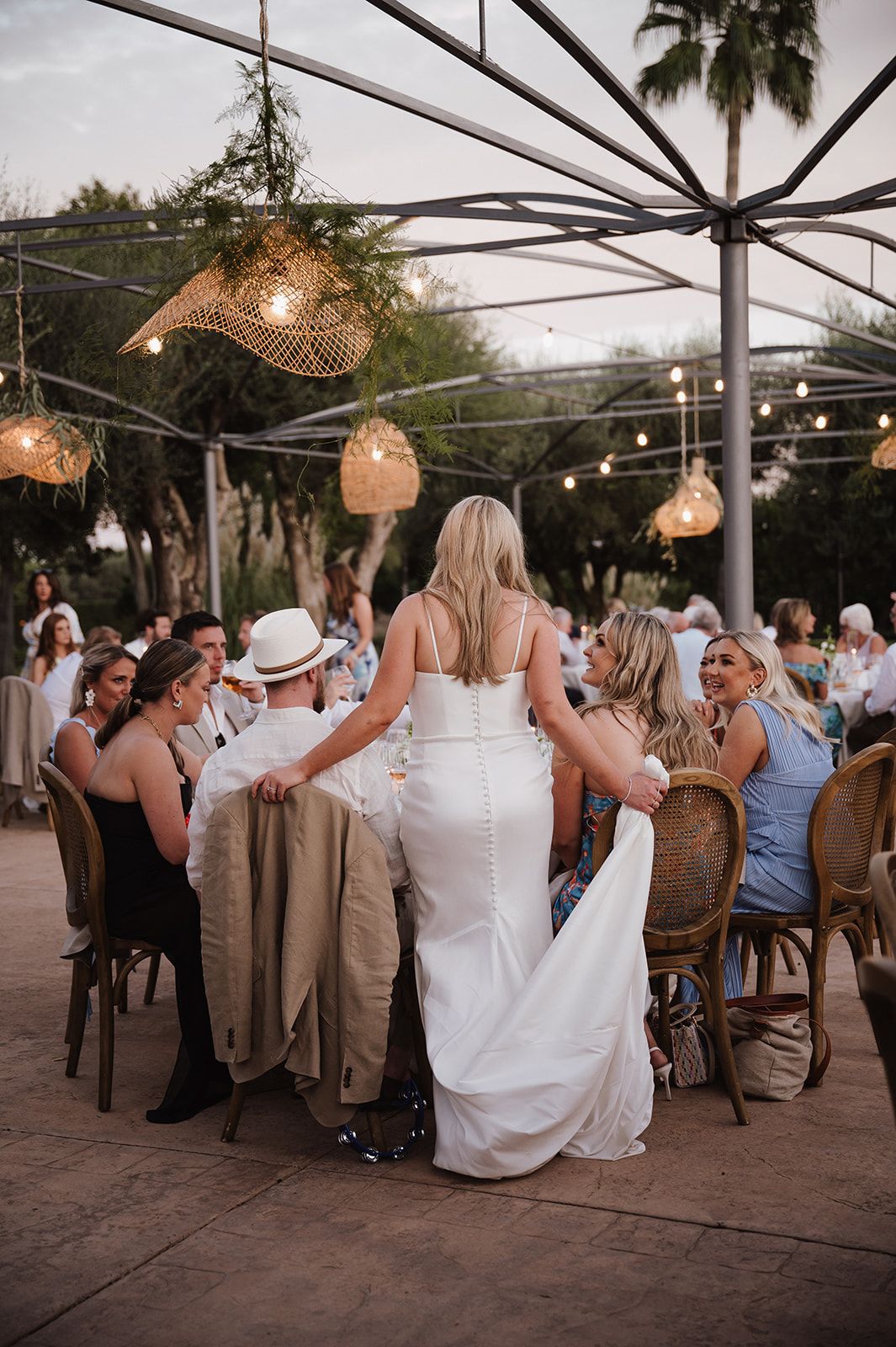 Bride in fitted white gown greets seated guests at outdoor wedding reception under string lights.