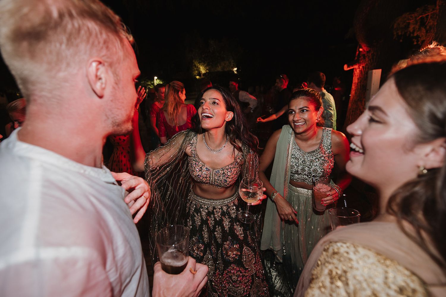 Bride in embroidered lehenga laughing with guests during outdoor Indian wedding reception celebration.