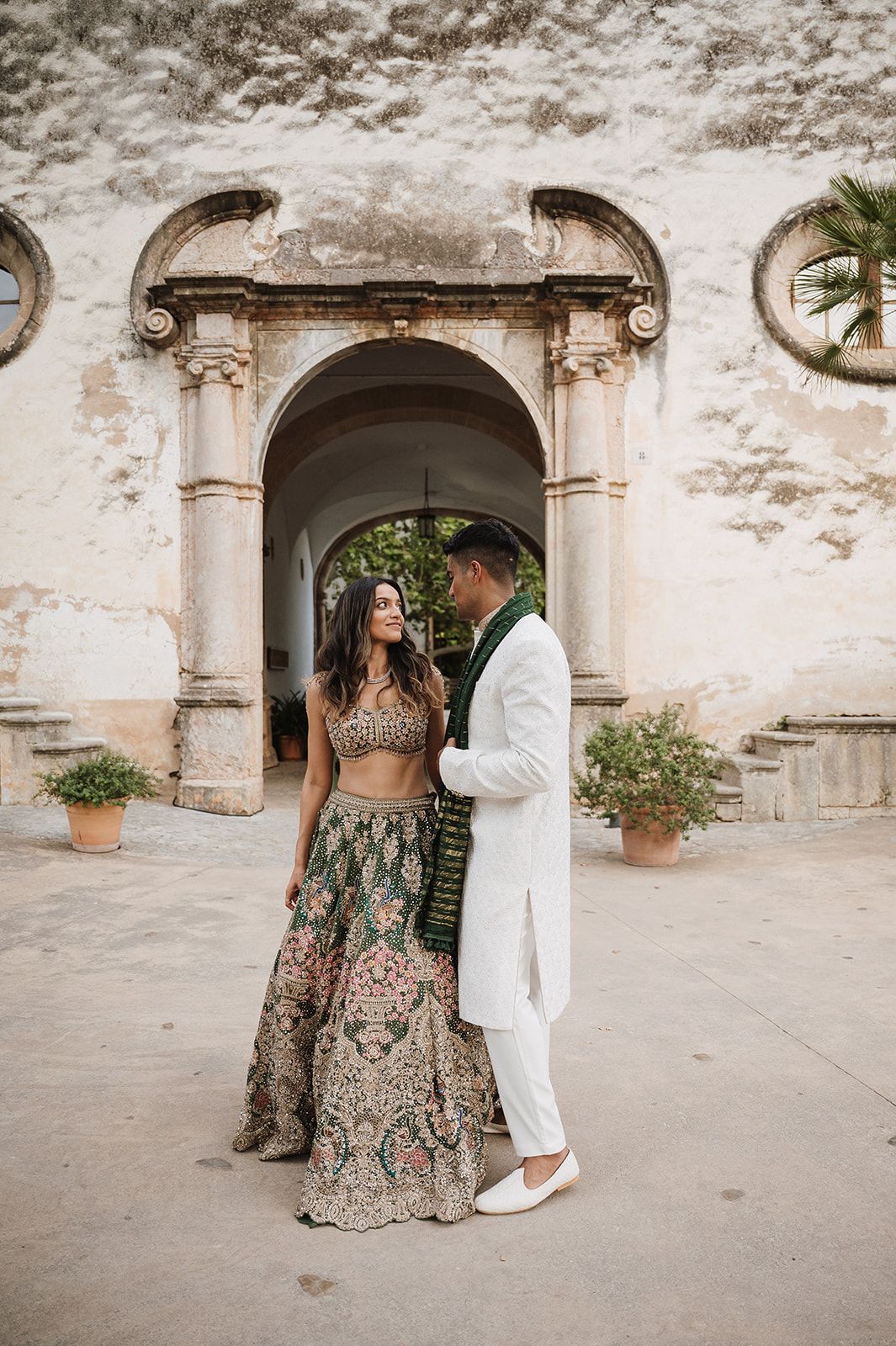 Bride in green embroidered lehenga and groom in white sherwani share a moment by a stone archway.