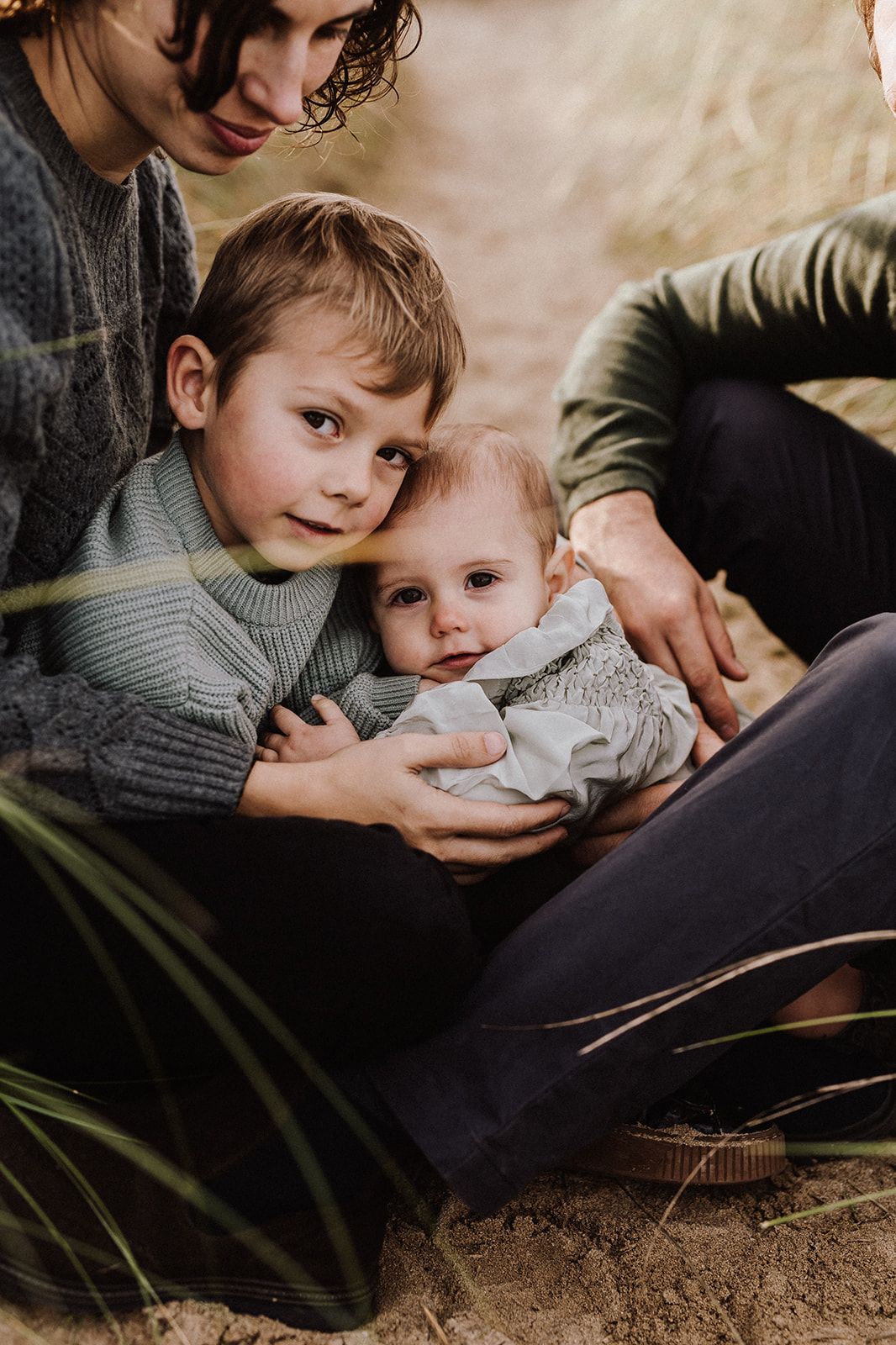 A young boy tenderly hugs a baby sibling while sitting outdoors with a parent in a warm, natural setting.