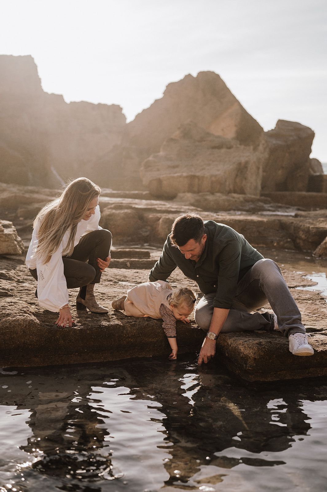 Family with young child exploring rocky tide pools together in golden sunlight near coastal cliffs.