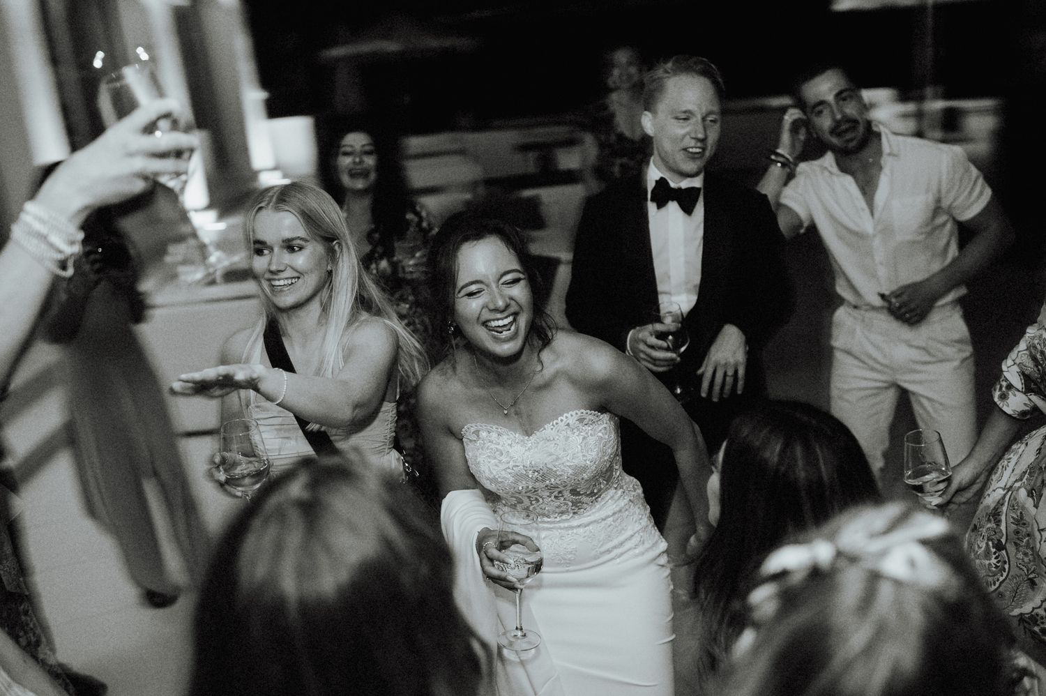 Black and white photo of bride laughing and dancing joyfully with guests at wedding reception.