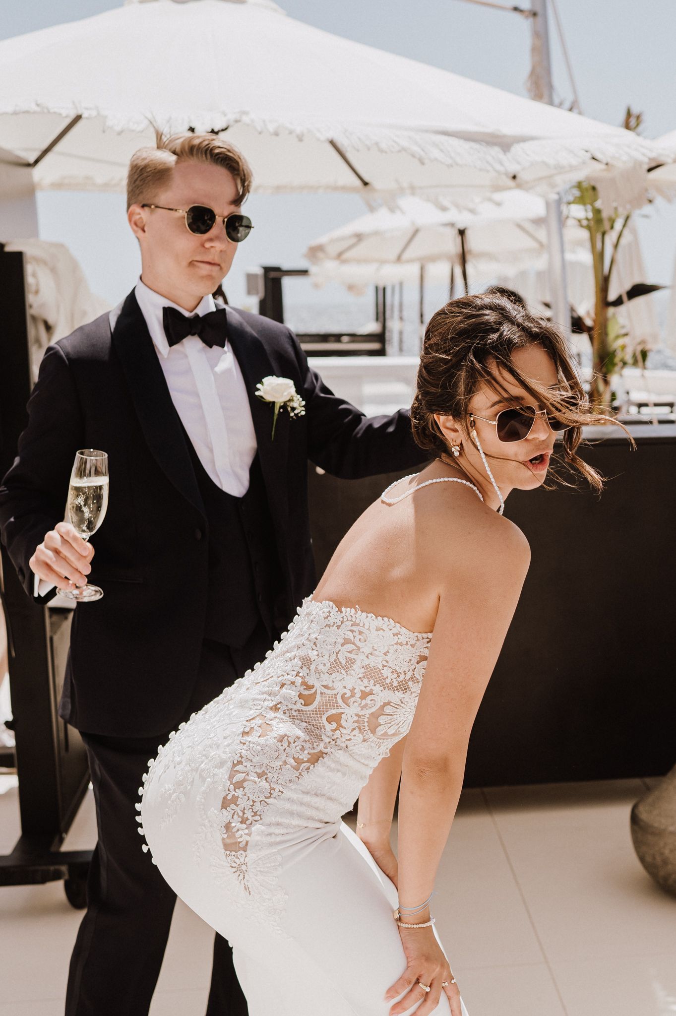Stylish bride in backless lace wedding dress and groom in black tuxedo holding champagne on rooftop.