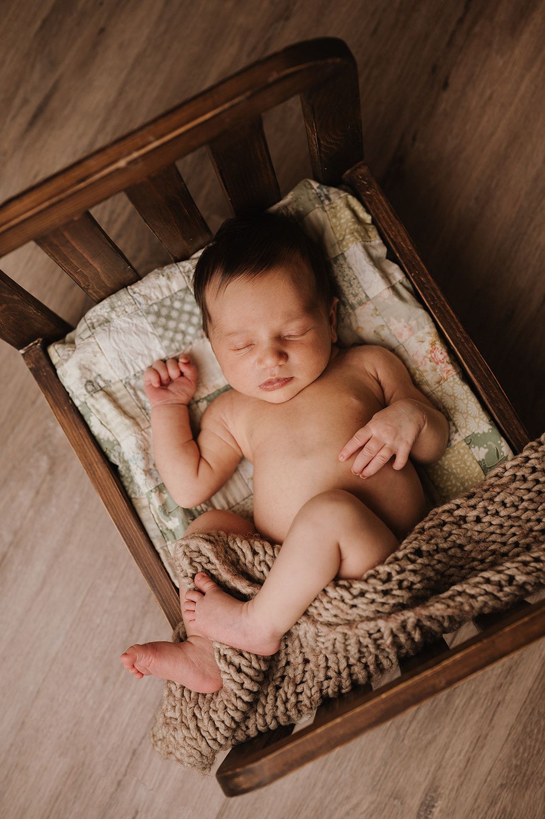 Peaceful newborn baby sleeping in a rustic wooden miniature bed with a knit blanket and floral quilt.