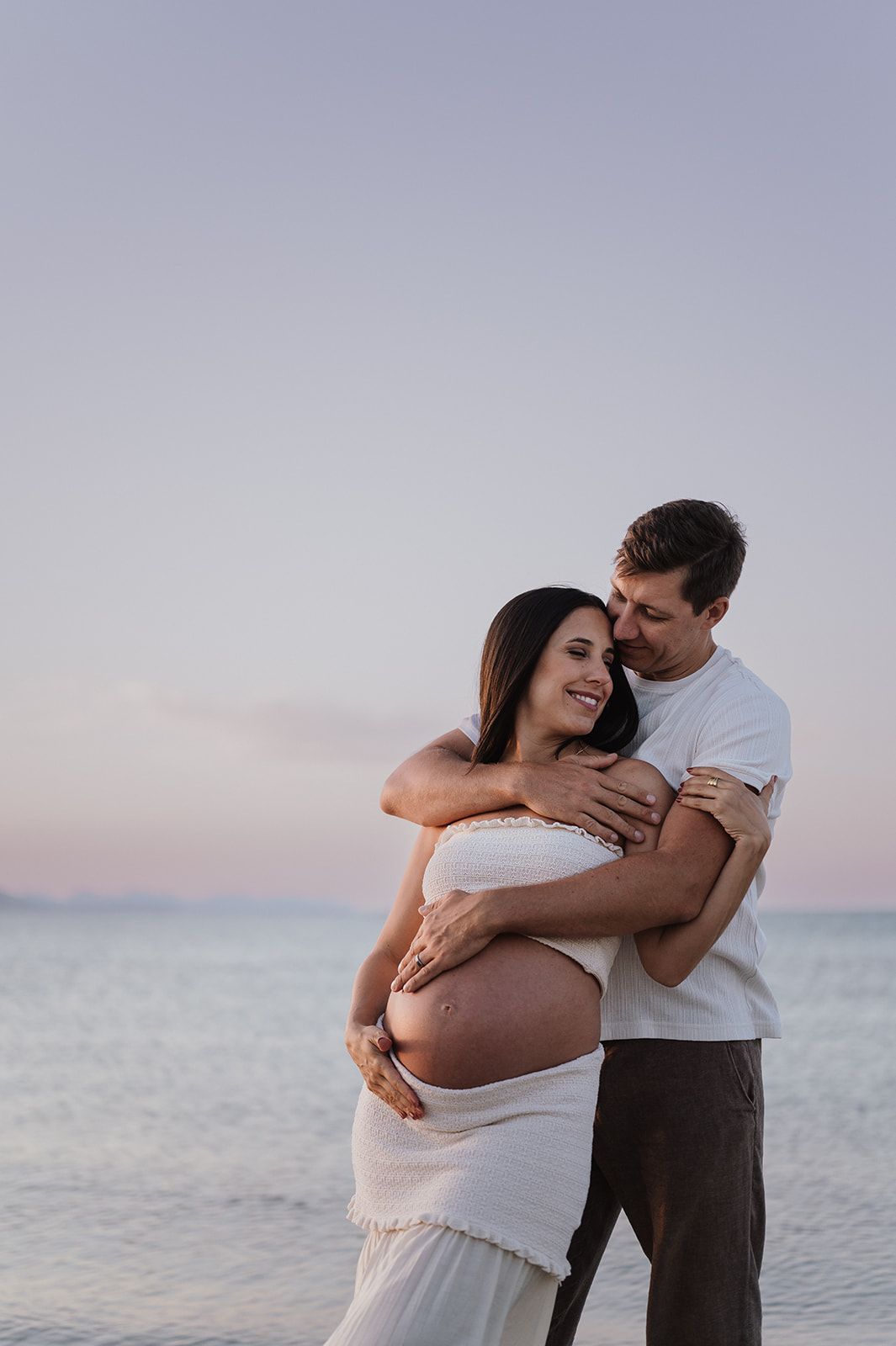Man tenderly embracing pregnant woman in white dress on beach, both smiling at sunset over calm water.
