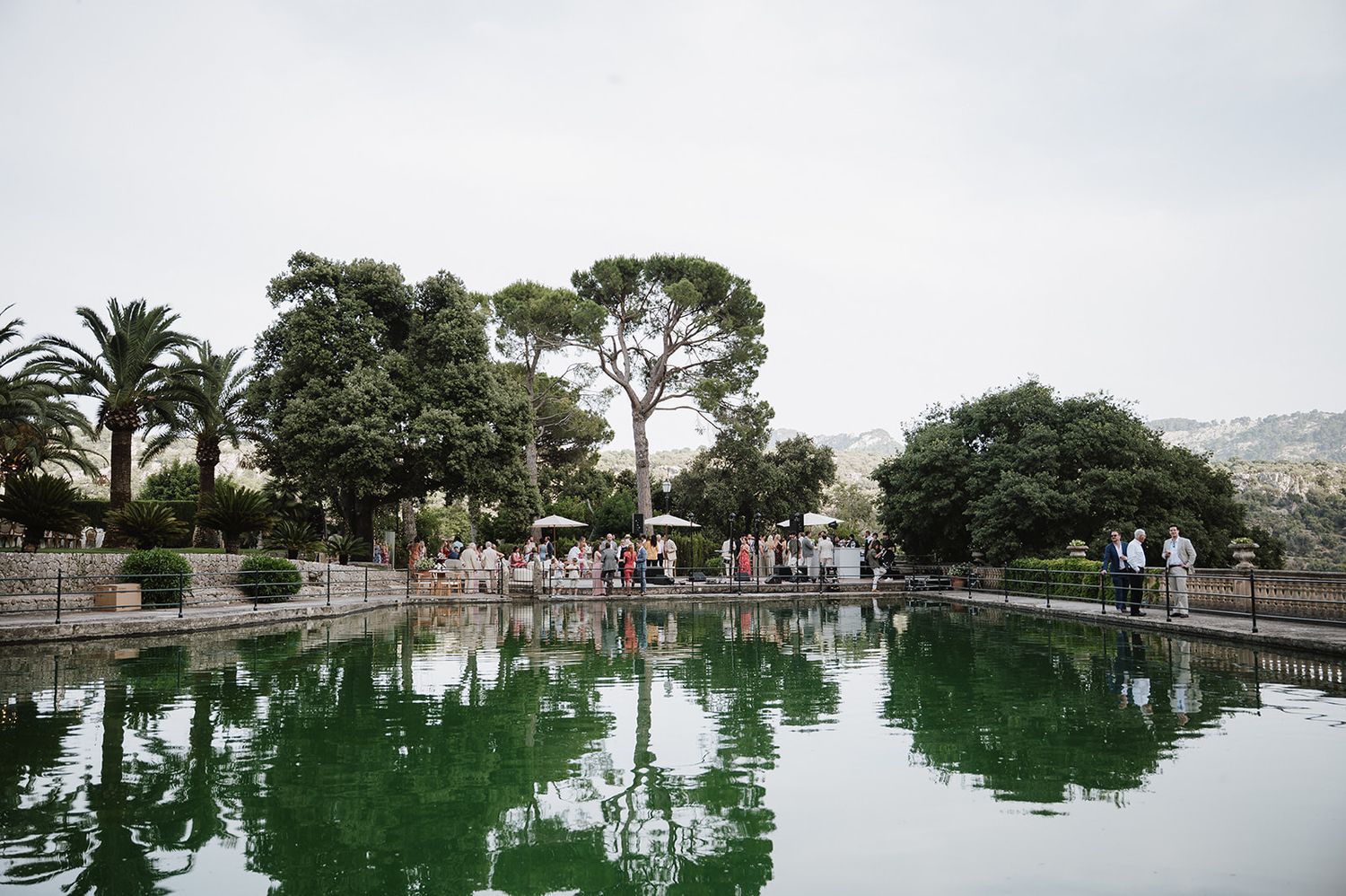 Guests gather near a serene reflective pond surrounded by lush trees at an elegant outdoor wedding venue.