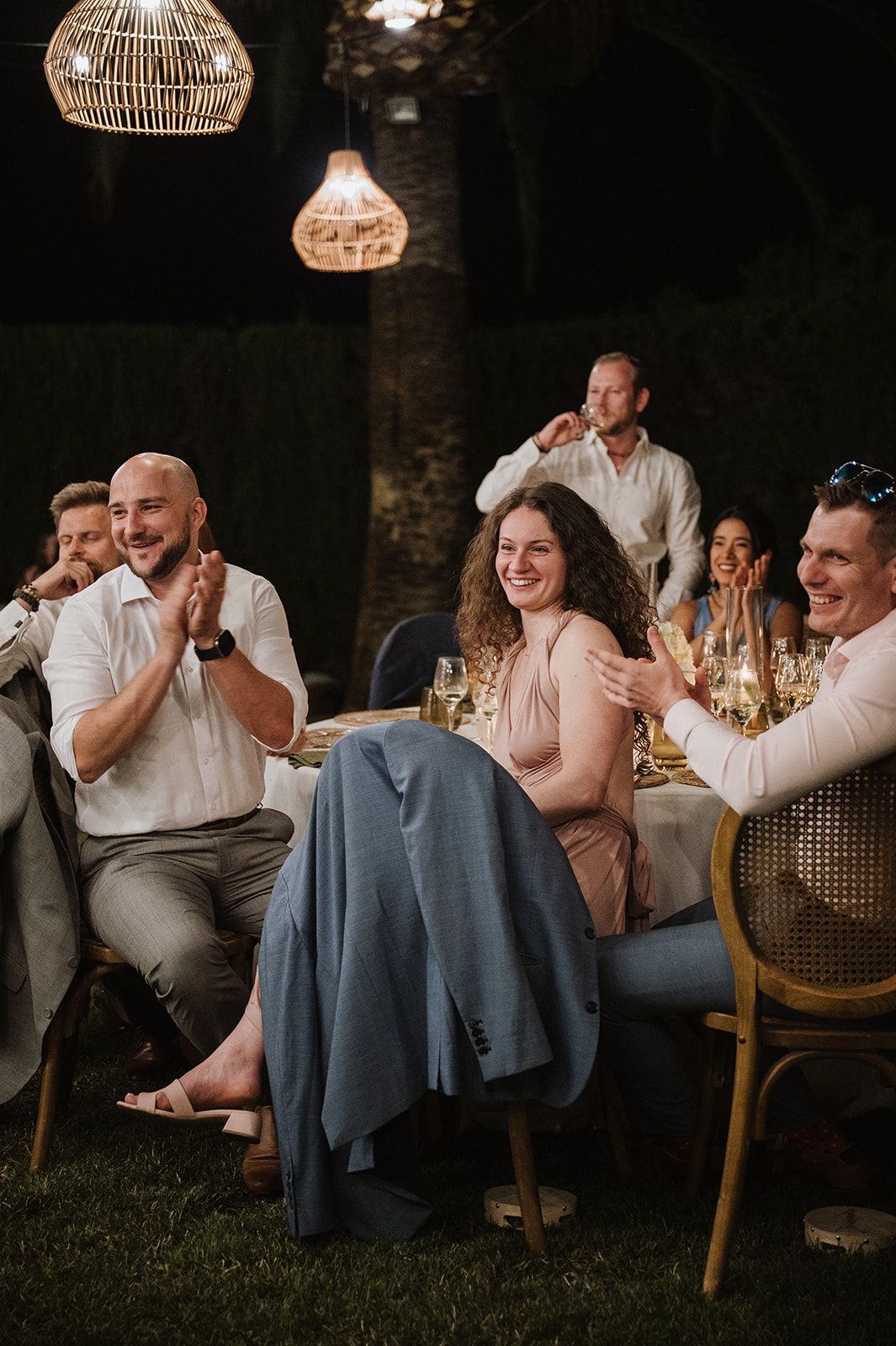 Wedding guests laugh and applaud during speeches at an outdoor evening reception under wicker pendant lights.