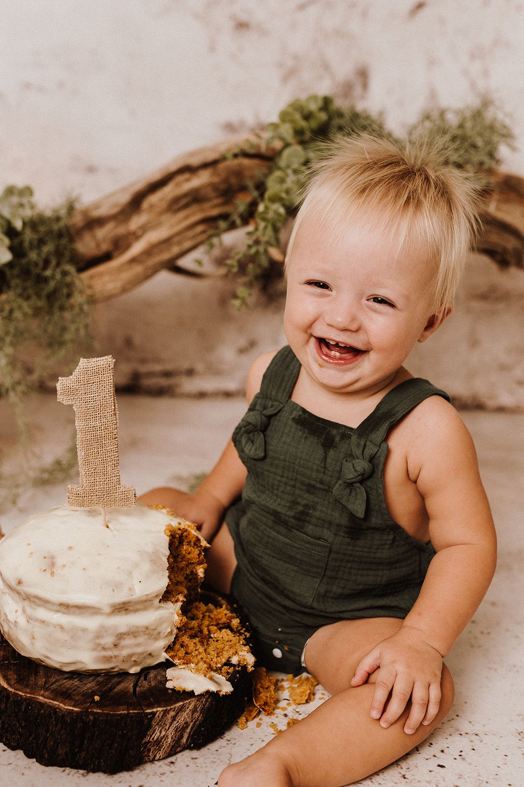 Smiling baby in green romper celebrates first birthday with smash cake topped with burlap number one candle.