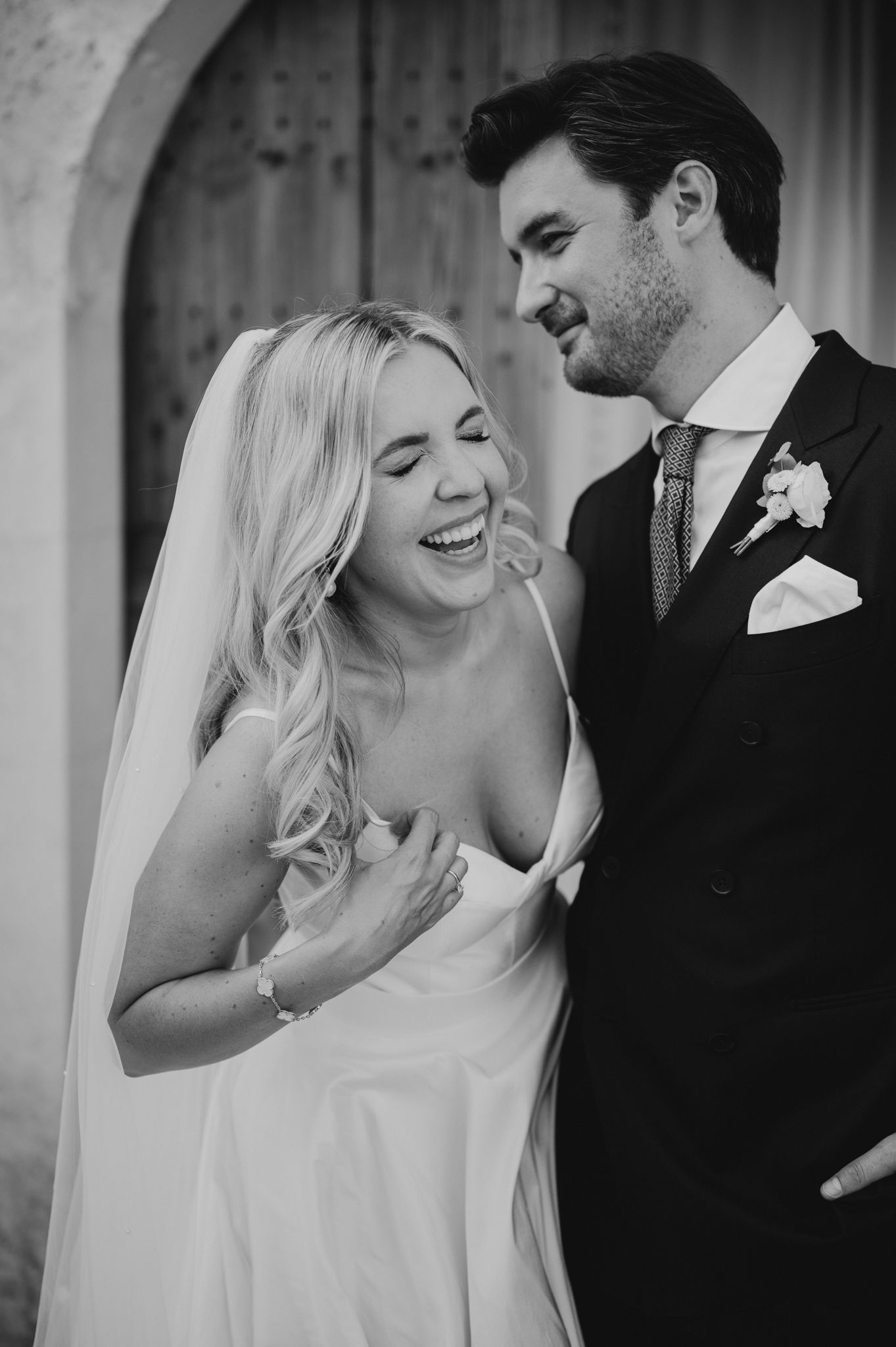 Black and white portrait of laughing bride in wedding gown with groom in dark suit at outdoor venue.