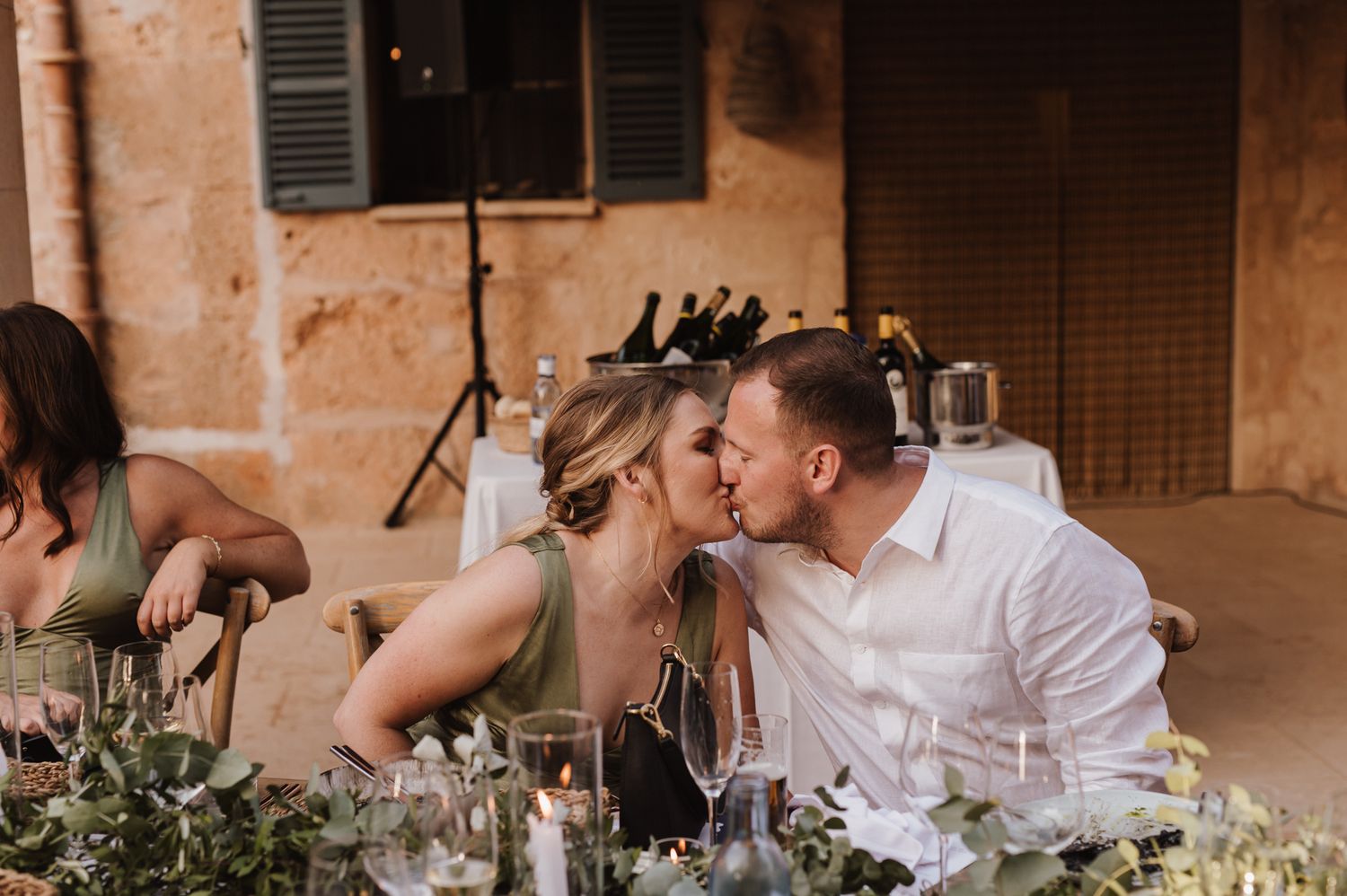 Couple shares a kiss at a candlelit wedding reception table decorated with lush greenery.
