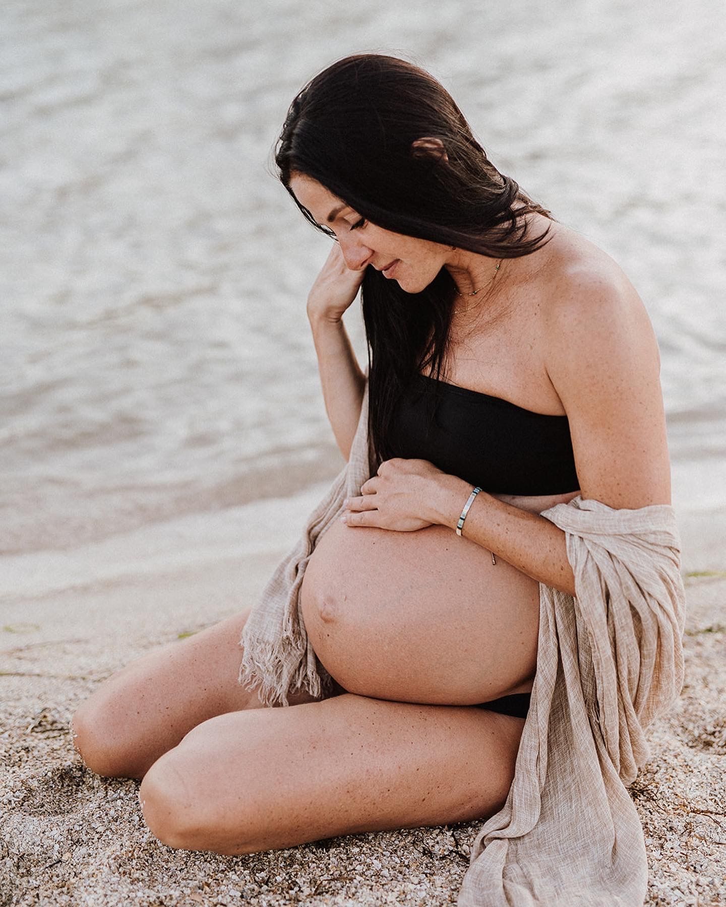 Pregnant woman sitting on sandy beach in black bikini top and beige wrap, cradling her baby bump near the water.