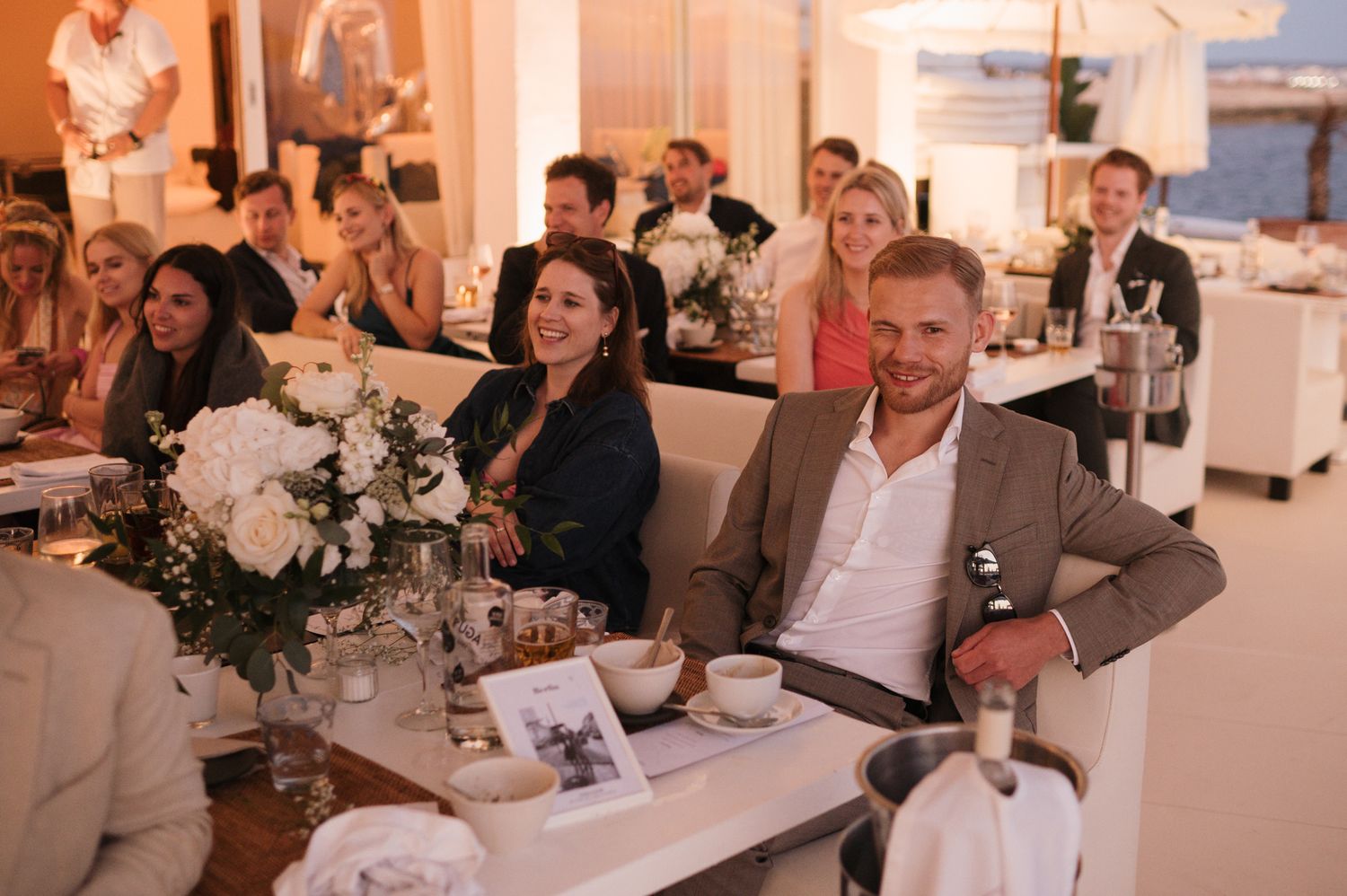 Wedding guests smile and laugh at elegant reception dinner with white floral centerpieces.