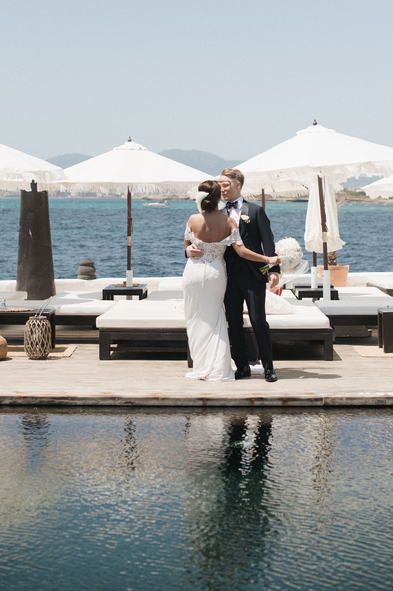 Bride and groom share a kiss beside a pool with ocean and white umbrellas in background.