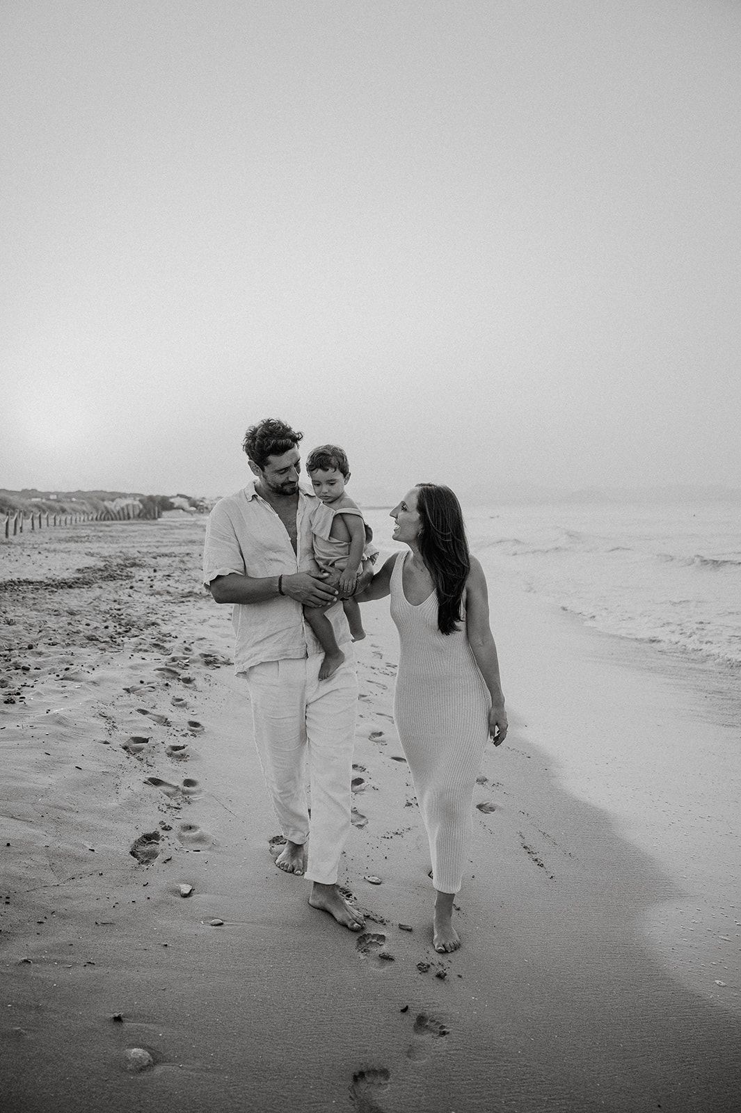 Black and white photo of a happy family walking on the beach, parents holding their toddler near ocean waves.