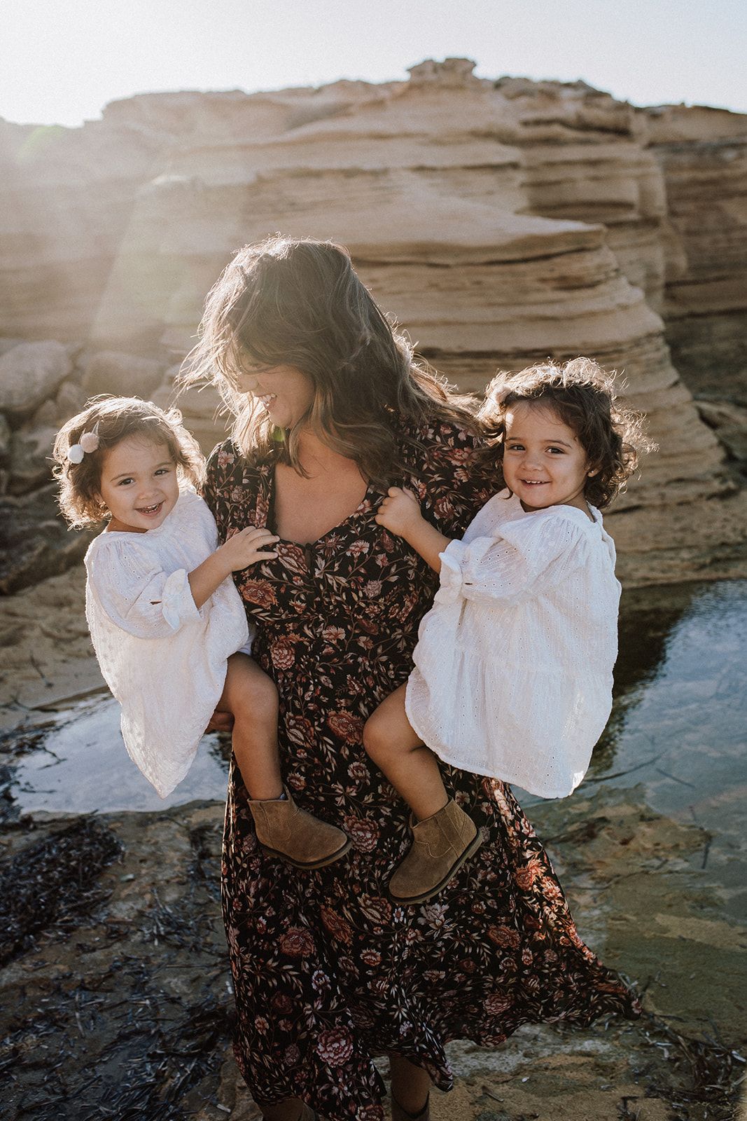 Mother holding two toddlers in white dresses on sunlit rocky coastal cliffs, all smiling joyfully.