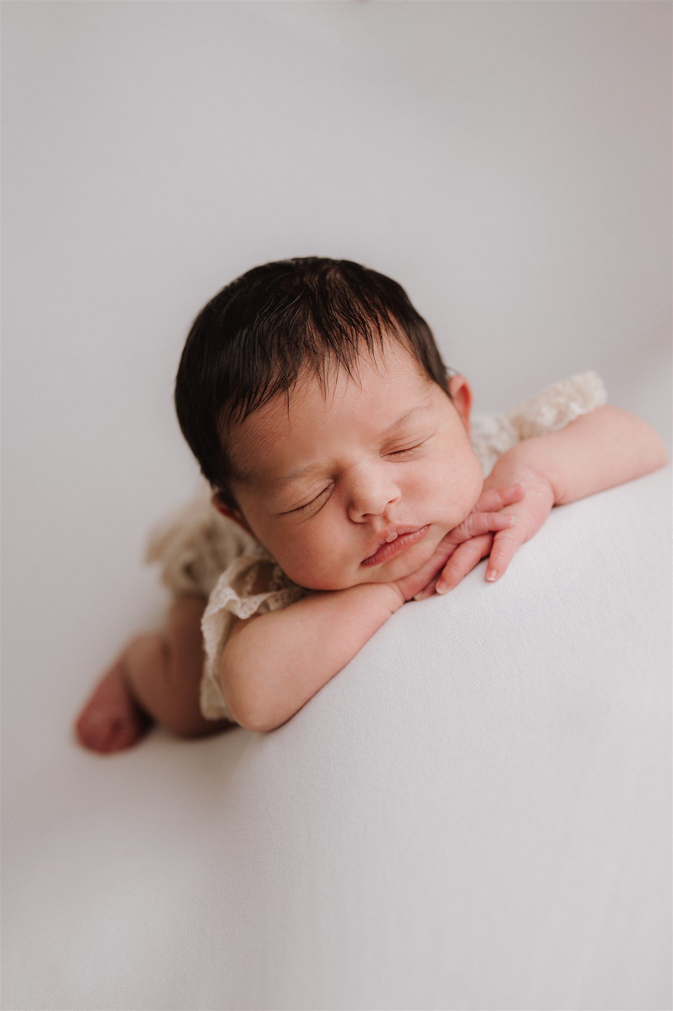 Peaceful newborn baby sleeping with hands tucked under chin, wearing cream romper on white background.