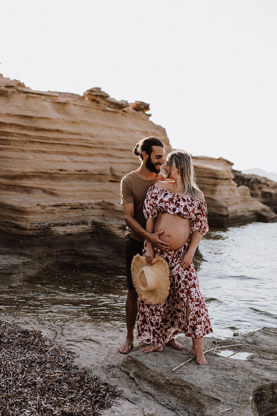 Couple sharing a tender moment on rocky beach, pregnant woman in floral dress holding straw hat.