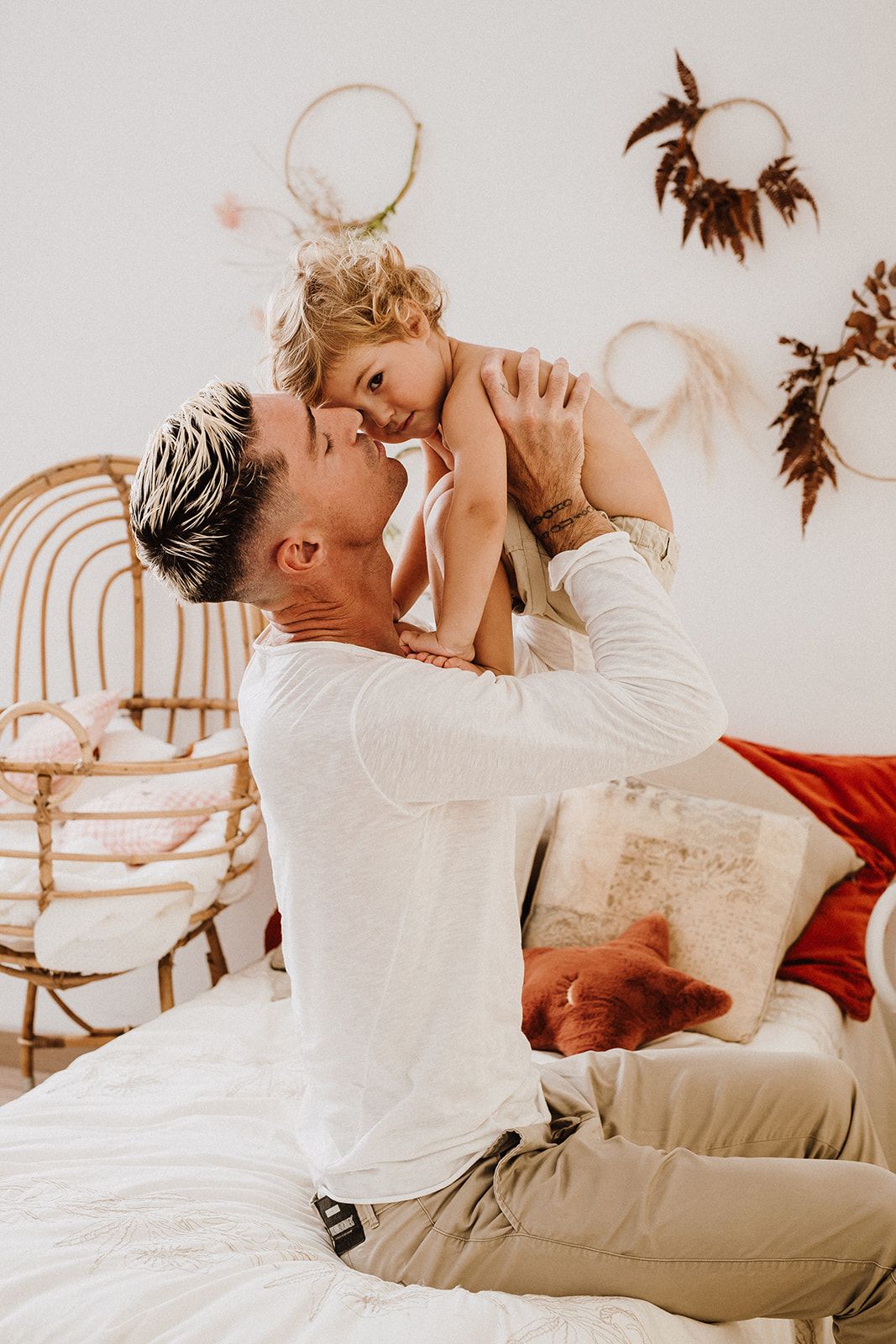 A father sits on a bed lifting his laughing toddler in a bright boho-styled bedroom with rattan decor.