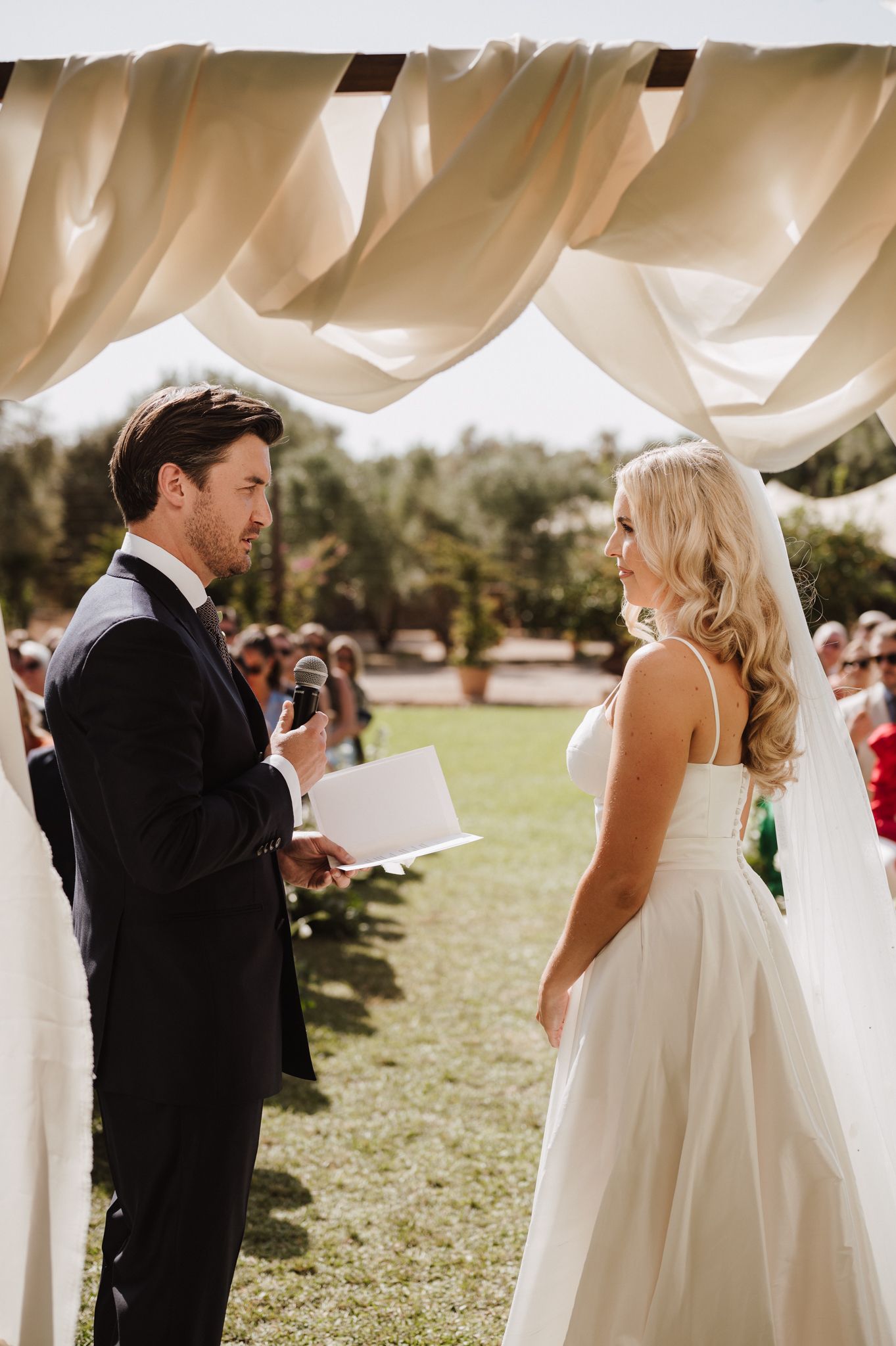Groom reading vows to bride under draped cream canopy at elegant outdoor wedding ceremony.