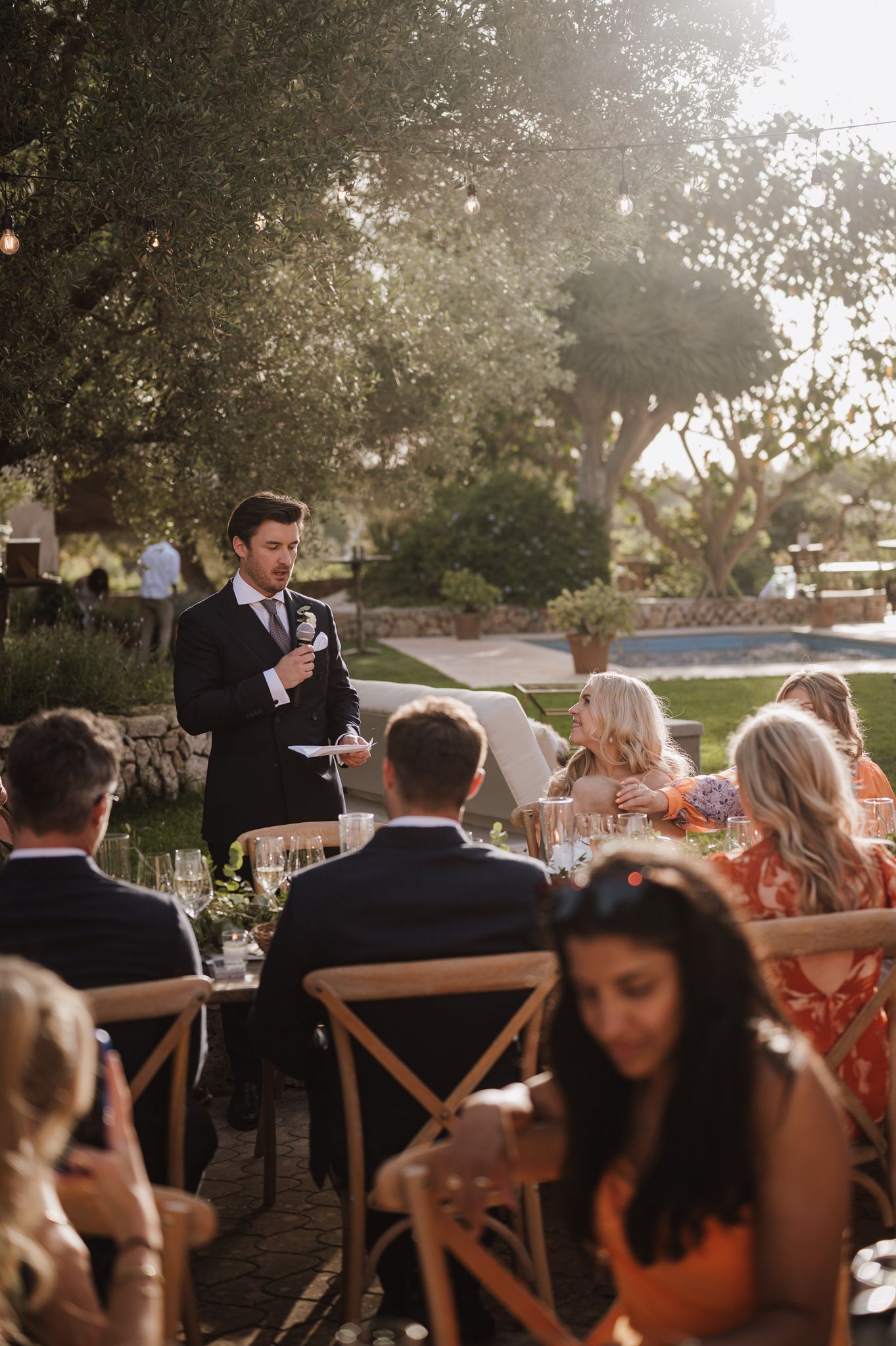 Groom giving speech at sunlit outdoor wedding reception while guests listen among olive trees.