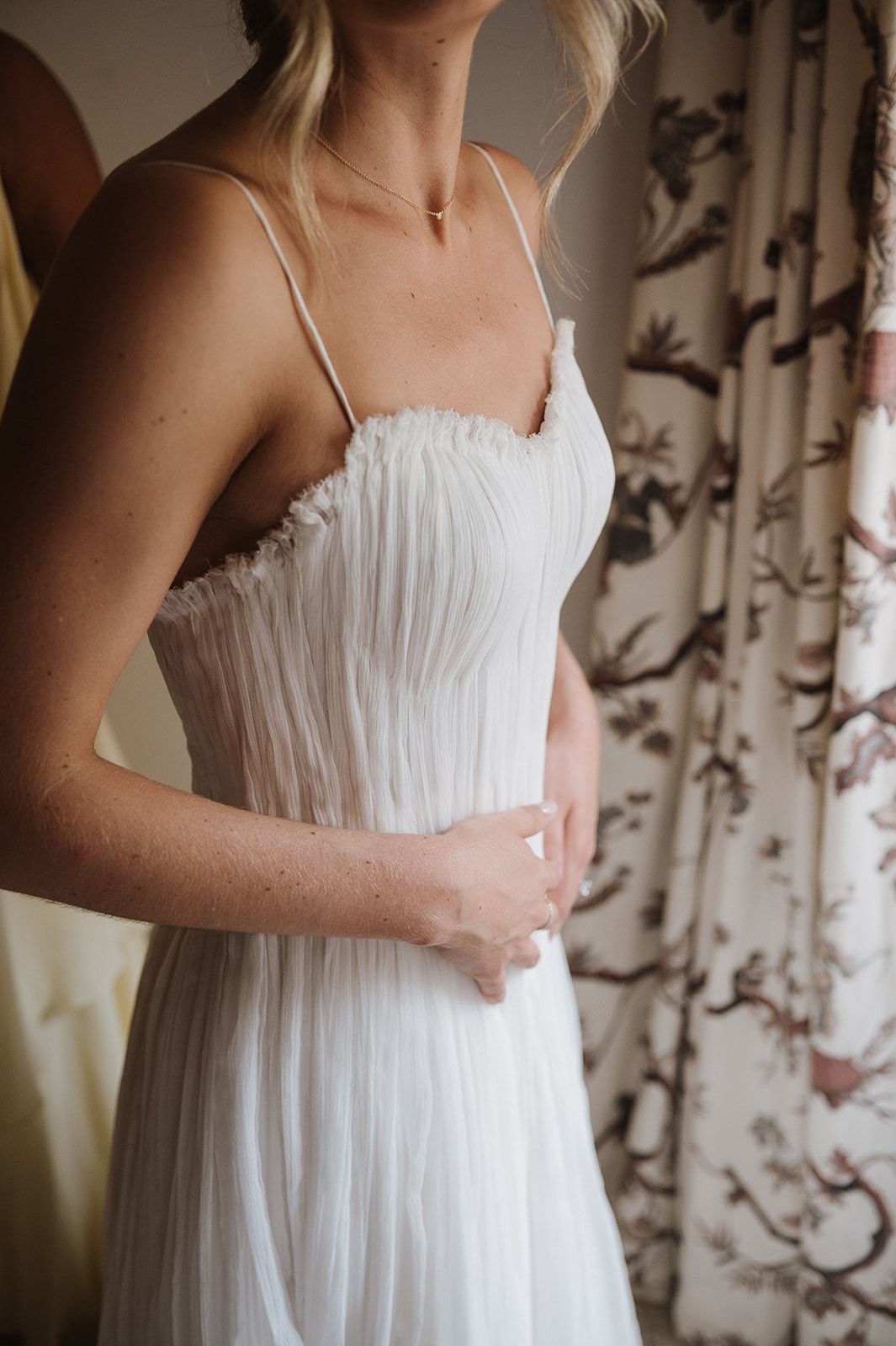 Bride in a white pleated tulle wedding gown with spaghetti straps and delicate ruffle bodice, standing near floral curtains.