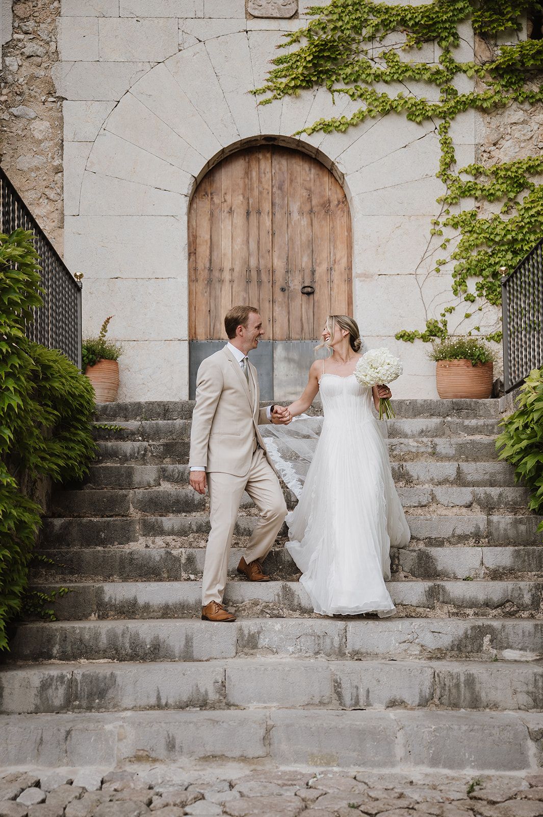 Bride and groom descend stone steps hand in hand before an ivy-covered arched wooden door.