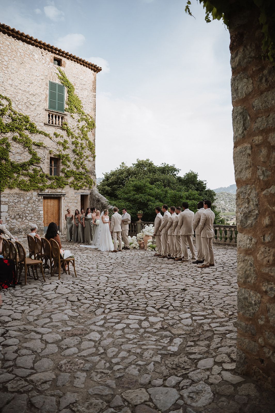 Wedding party stands on a rustic cobblestone courtyard beside an ivy-covered stone villa with mountain views.