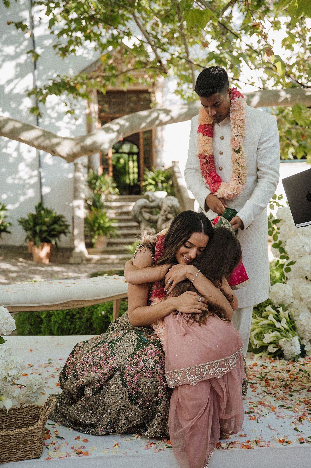Emotional Indian wedding moment as bride hugs guest while groom in white sherwani with floral garland looks on.