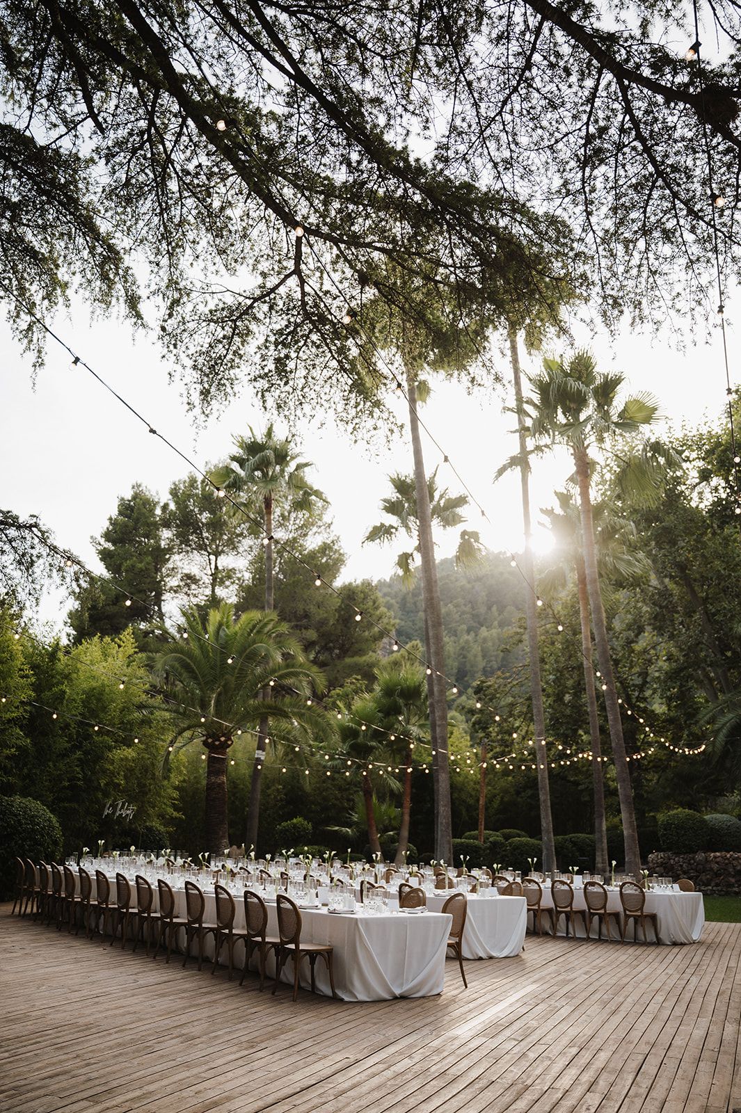Elegant outdoor wedding reception on a wooden deck with long white tables, string lights, and lush palm trees at sunset.