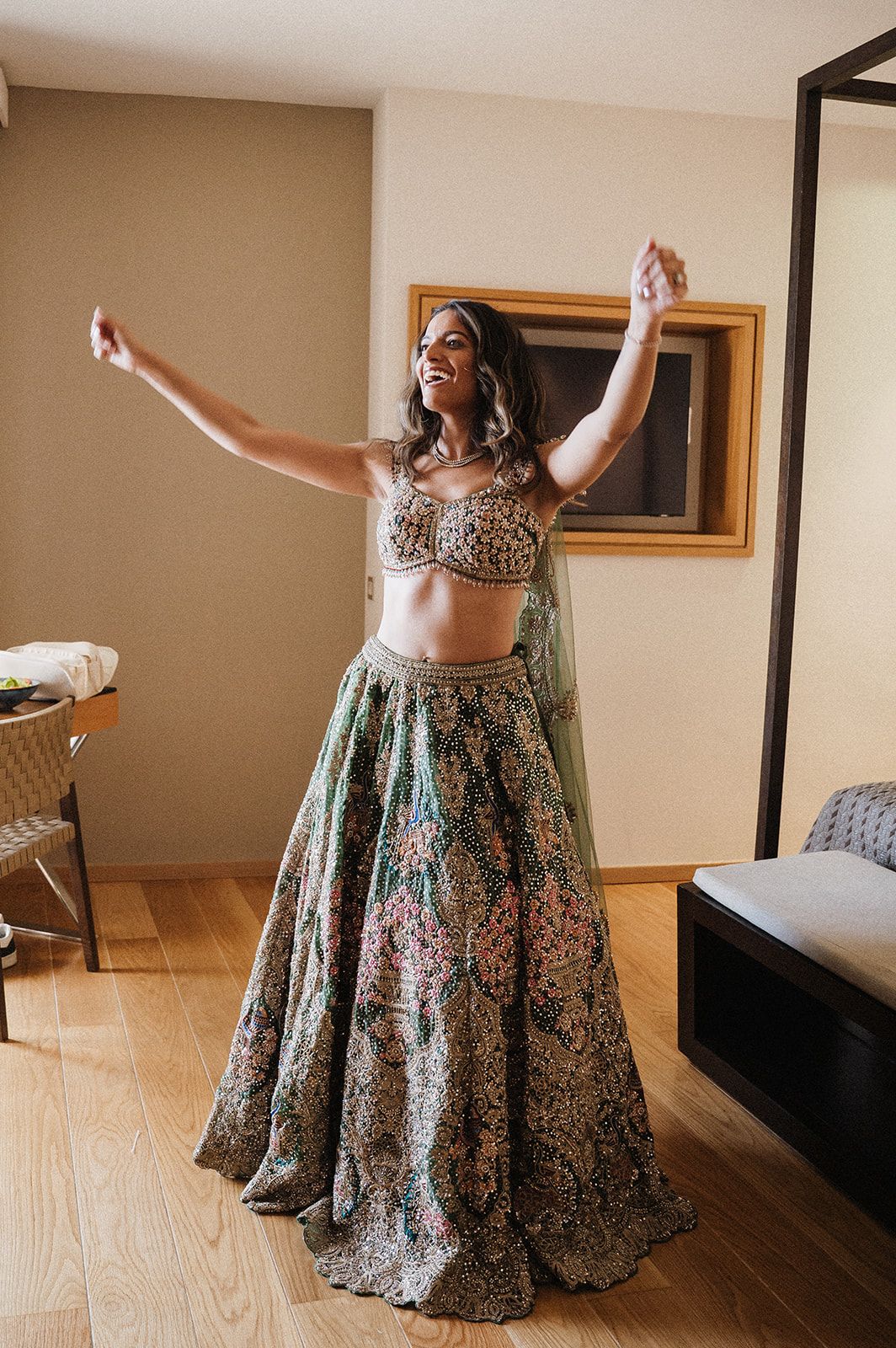 Bride joyfully twirling in a floral green lehenga choli with arms outstretched in a hotel room.