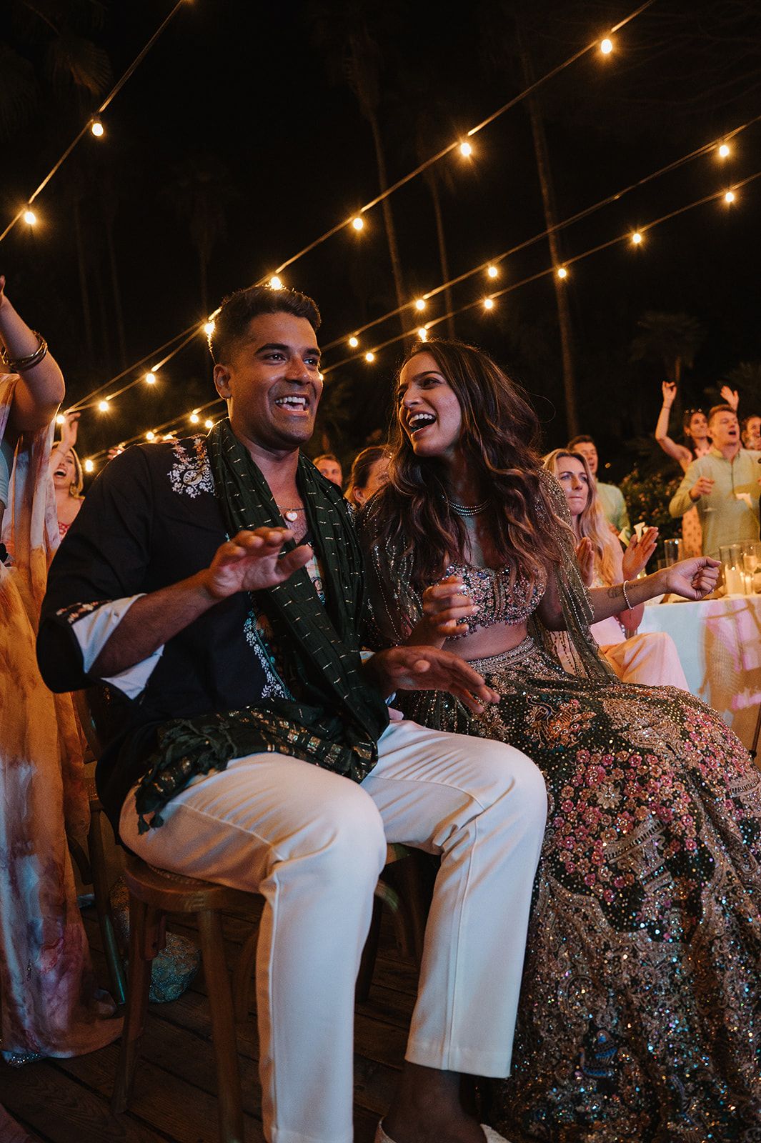 Bride and groom in Indian wedding attire laugh joyfully at outdoor evening reception.
