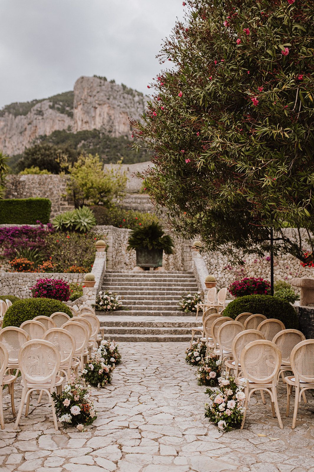 Outdoor wedding ceremony setup with rattan chairs and floral arrangements against dramatic mountain backdrop.