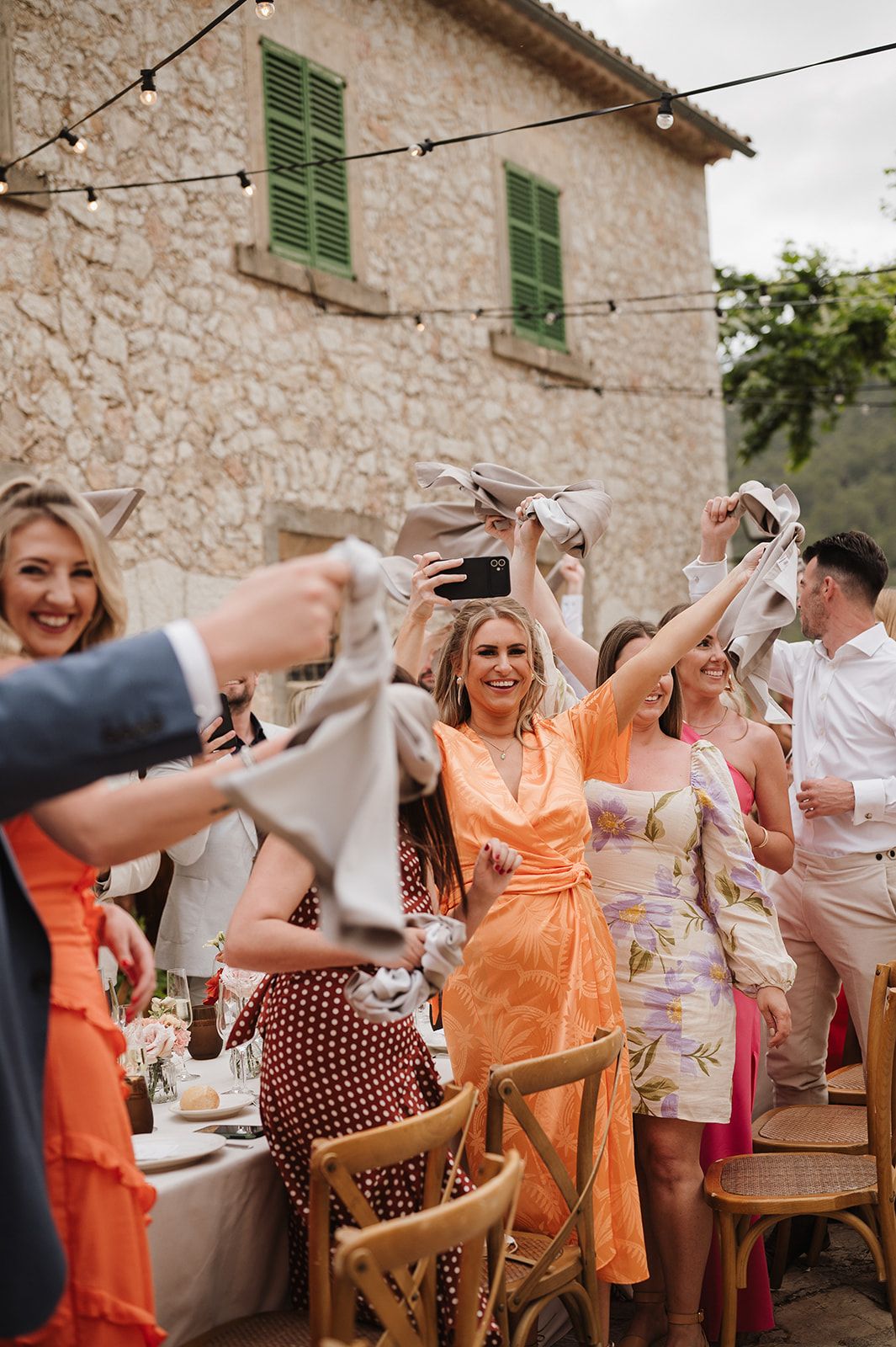 Wedding guests in colorful dresses joyfully waving napkins at an outdoor reception celebration.