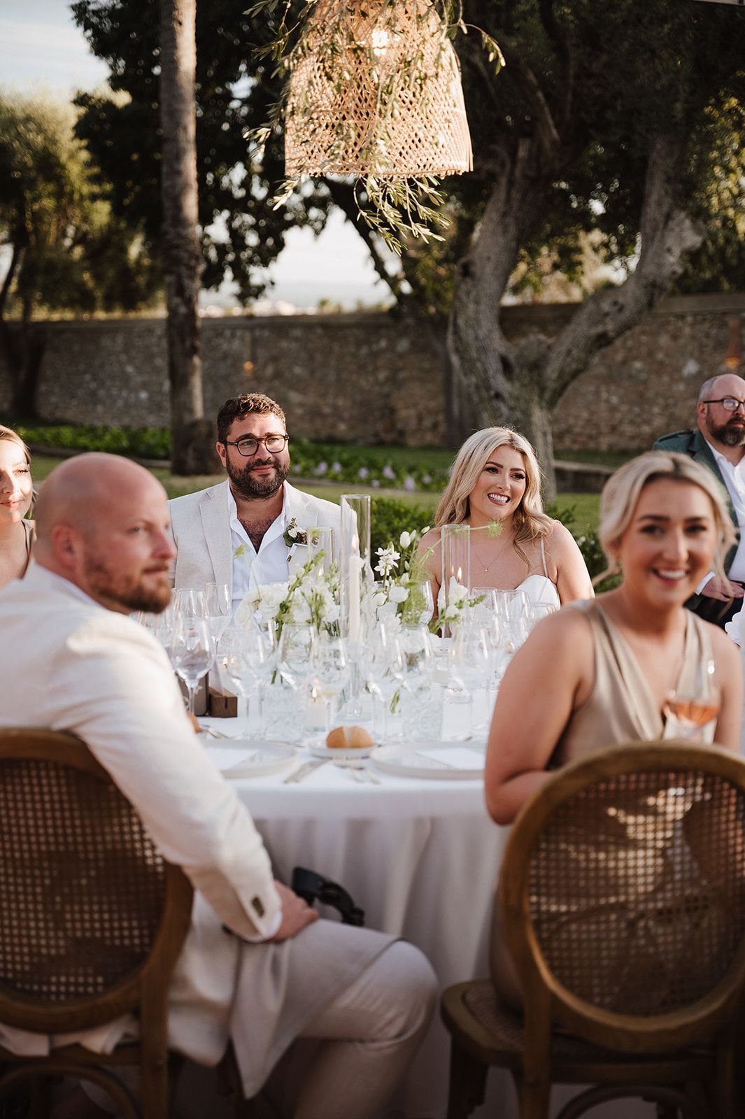 Bride and groom smile at outdoor wedding reception dinner table surrounded by guests at golden hour.
