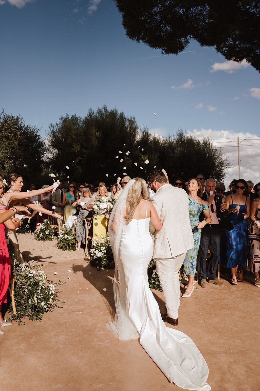 Newlyweds walk down the aisle as guests toss white flower petals in celebration outdoors.