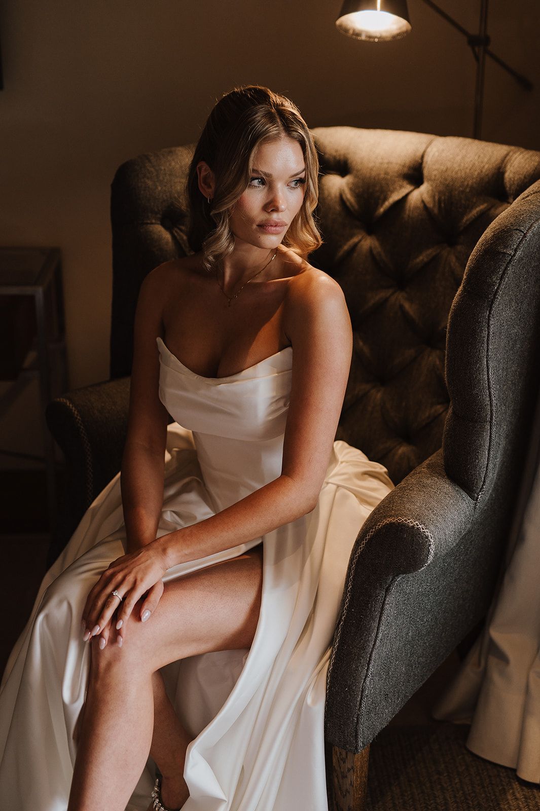 Elegant bride in strapless white satin gown sits gracefully in tufted gray velvet armchair in warm moody lighting.