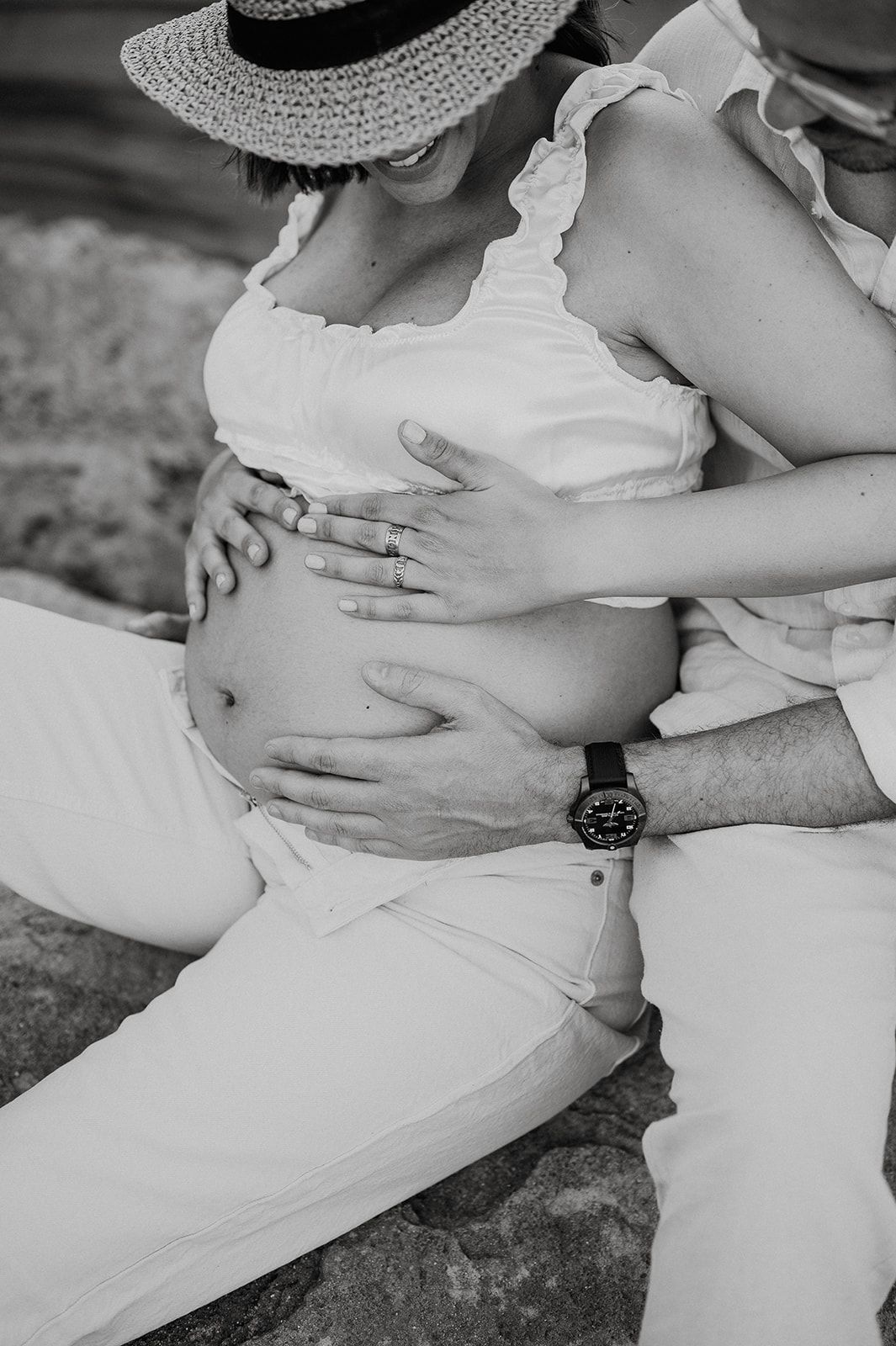 Black and white maternity photo of a couple cradling a pregnant belly, woman wearing a straw hat and white outfit.