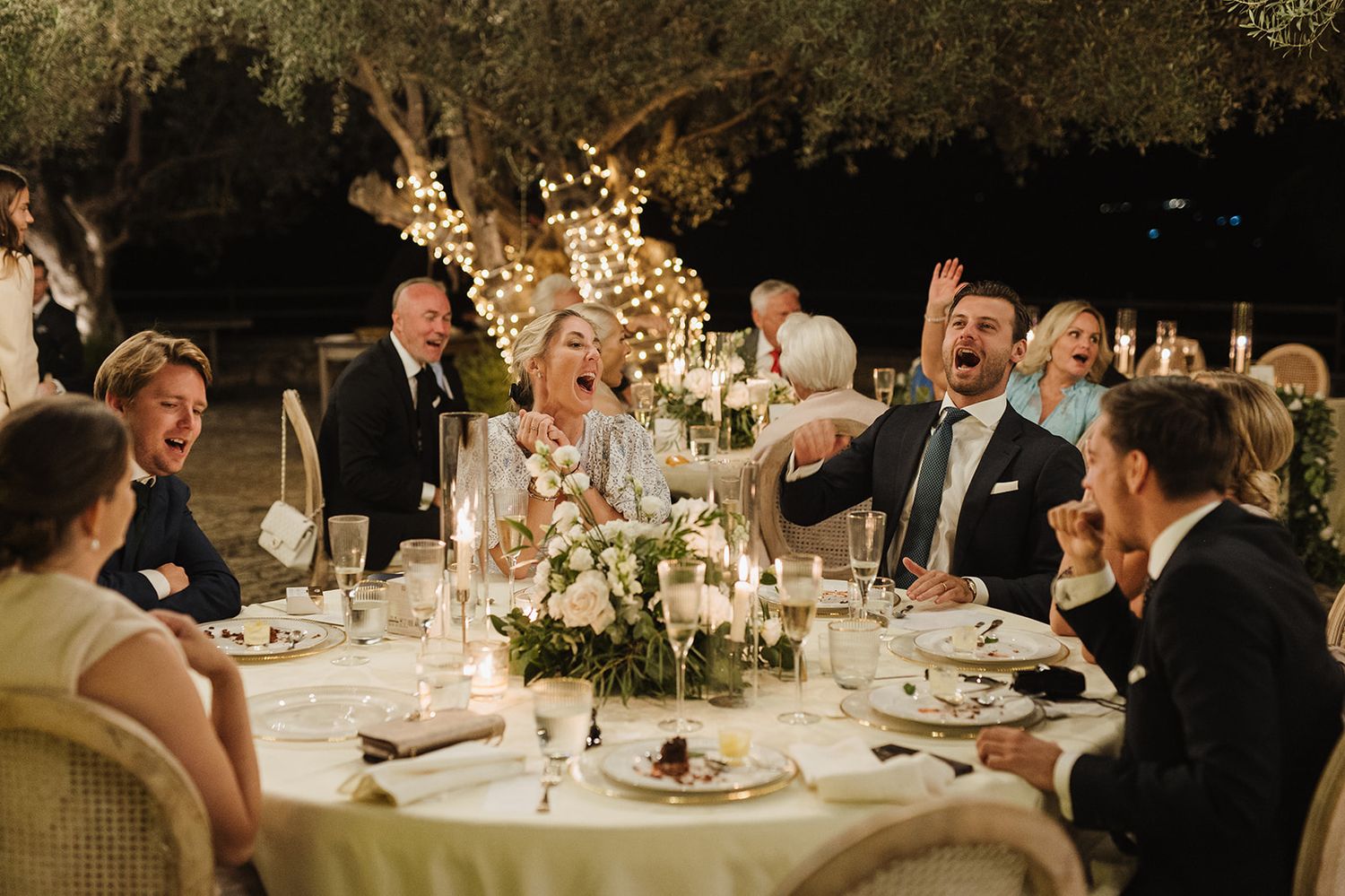 Wedding guests laughing and celebrating joyfully at a candlelit outdoor reception table at night.