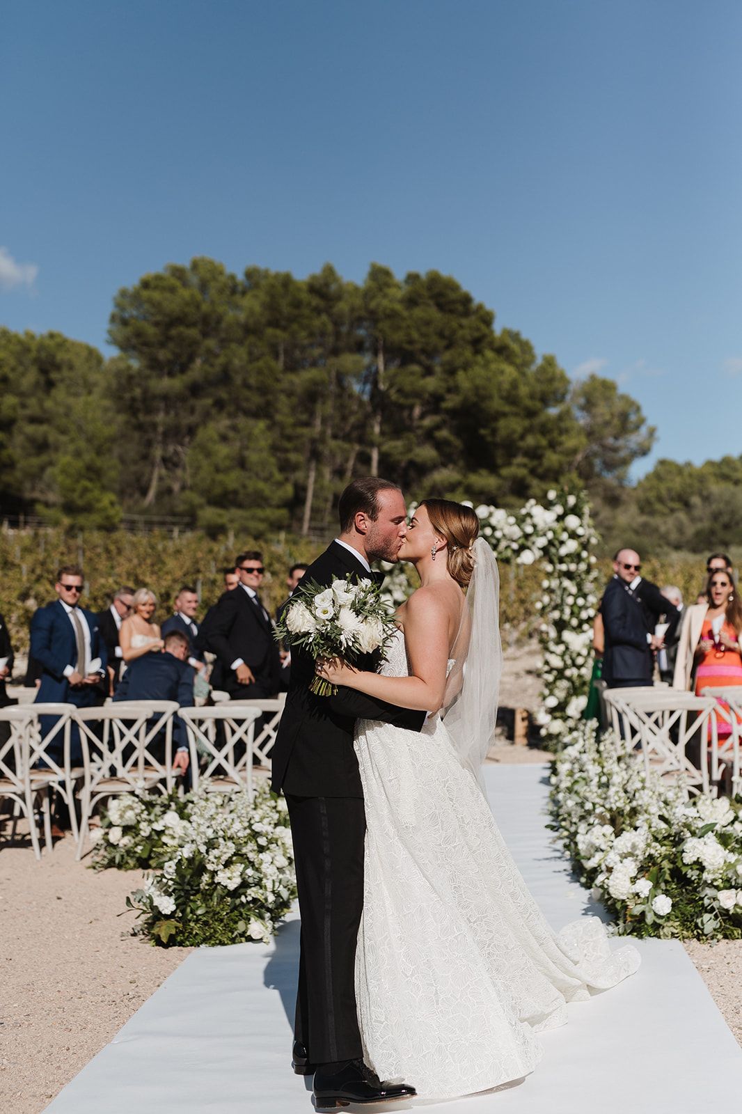 Bride and groom kissing at outdoor ceremony aisle with white floral arch.