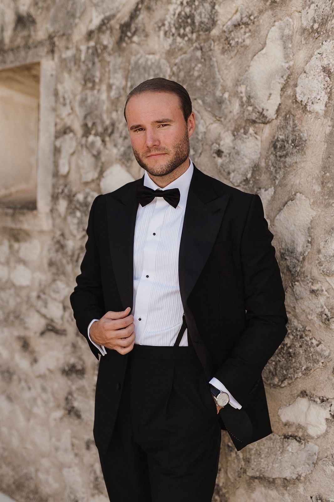 Groom in black tuxedo with bow tie posing against rustic stone wall.