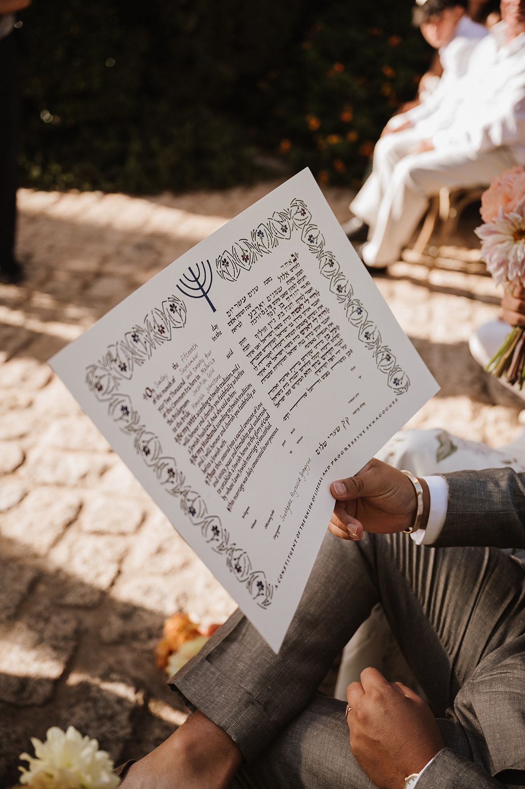 Guest holding a Jewish ketubah marriage contract with decorative border during outdoor ceremony.