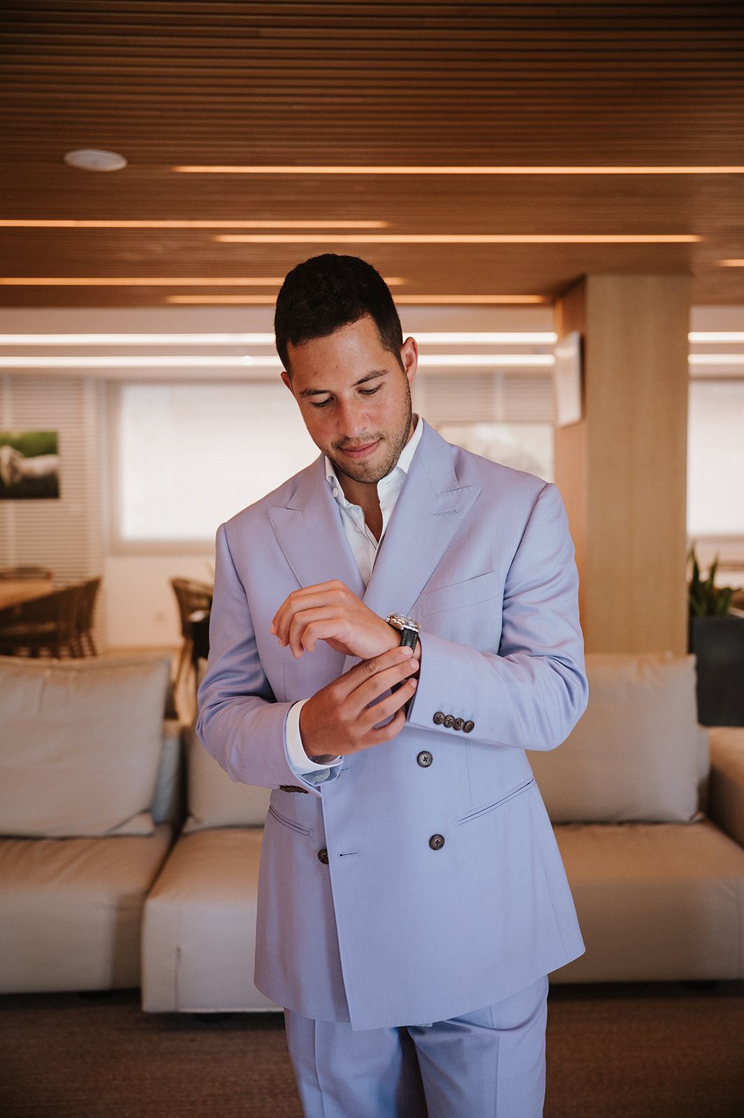 Groom in light blue double-breasted suit adjusting cufflinks indoors before wedding ceremony.