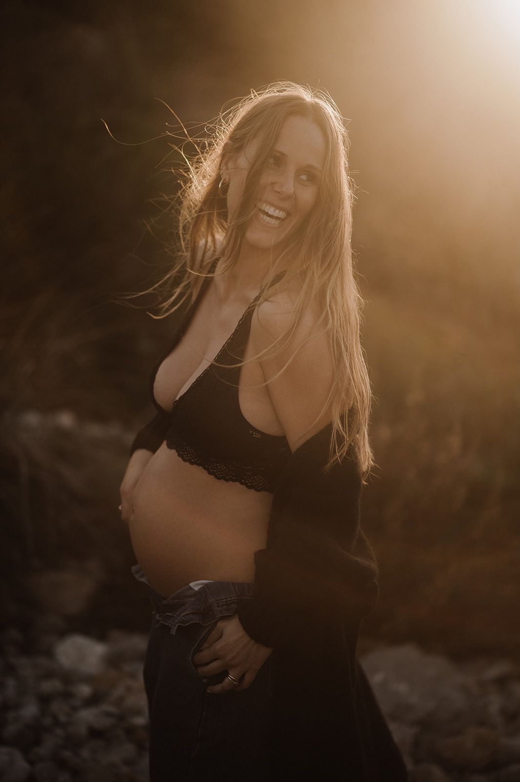 Smiling pregnant woman in black crop top posing outdoors in golden sunset light with wind in her hair.