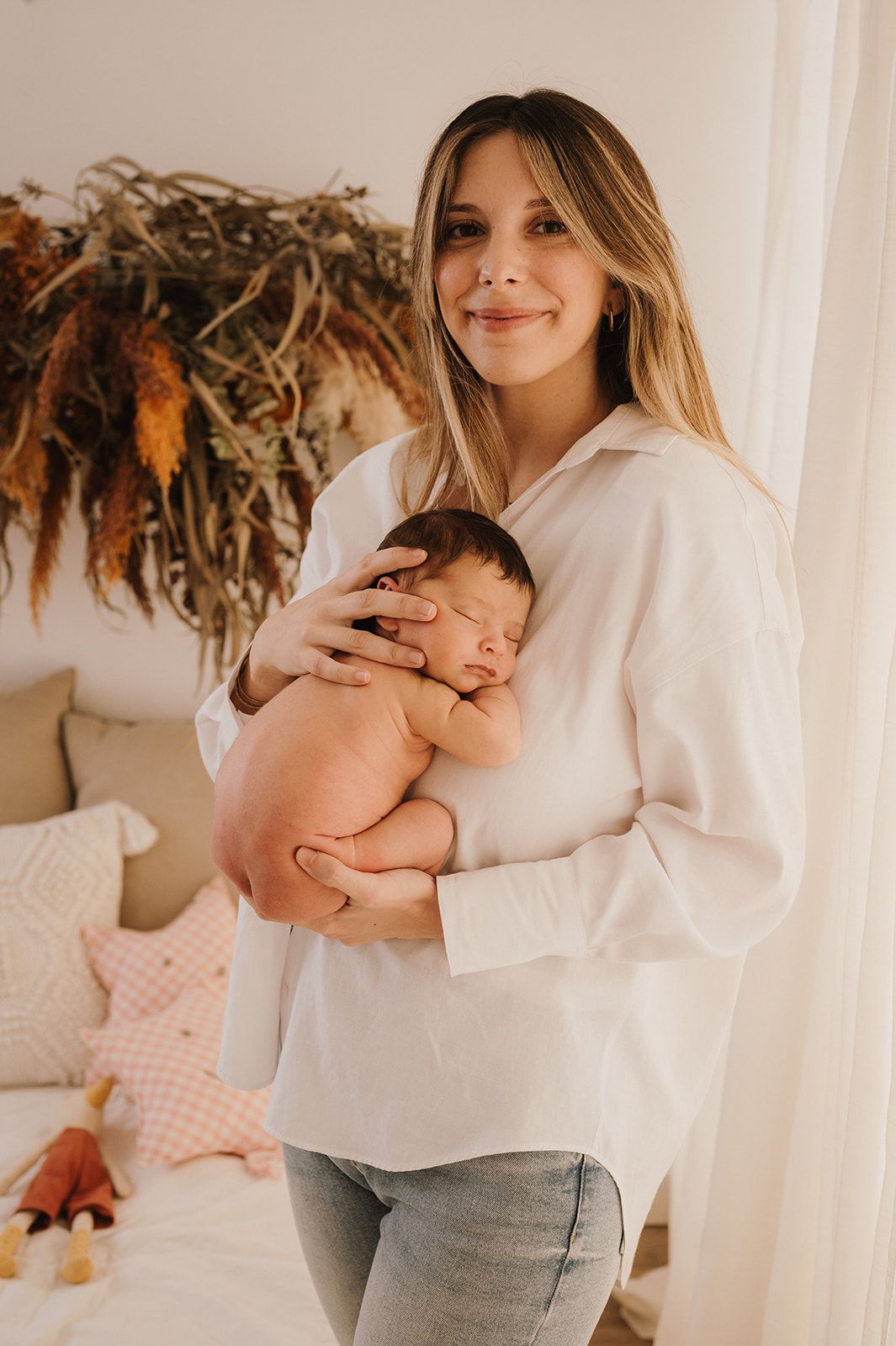 A smiling mother in a white shirt gently holds her sleeping newborn baby against her chest in a cozy room.