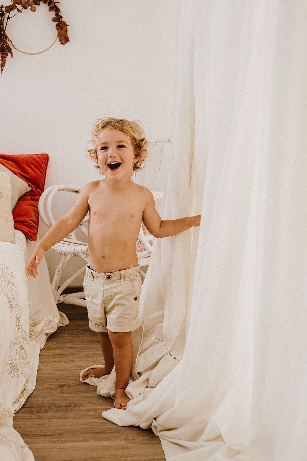A happy shirtless toddler boy laughs while playing with white curtains in a cozy boho bedroom.