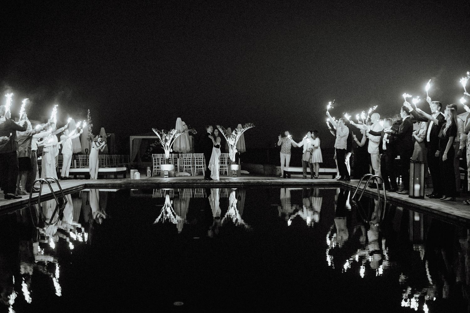 Wedding guests hold sparklers poolside at night, their reflections mirrored in the still water below.