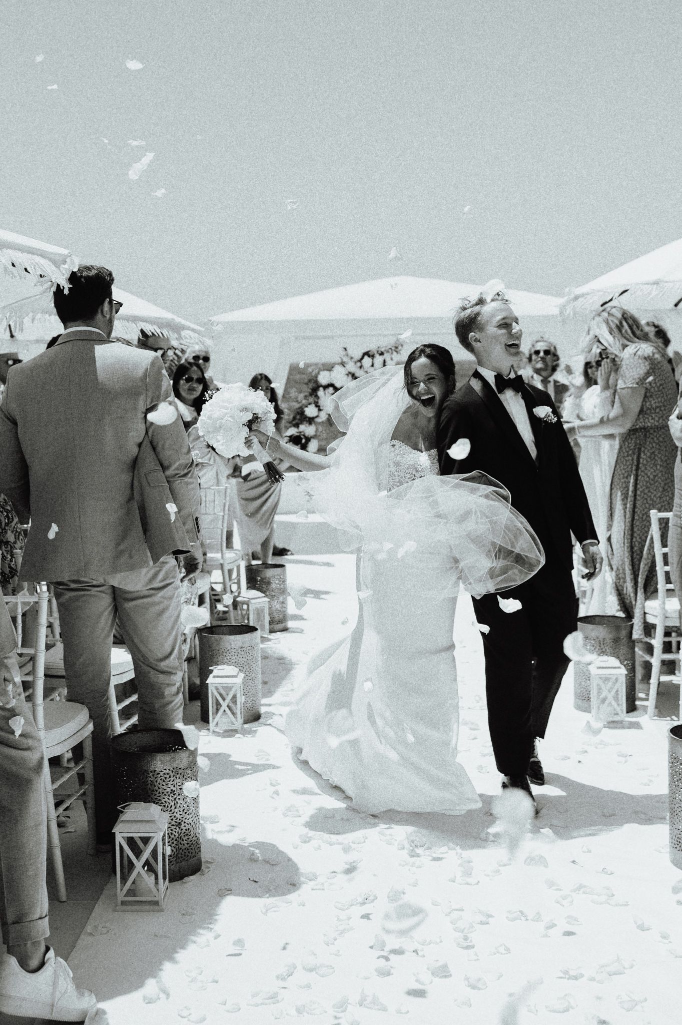 Black and white photo of joyful newlyweds walking down aisle as guests celebrate with confetti at beach wedding.