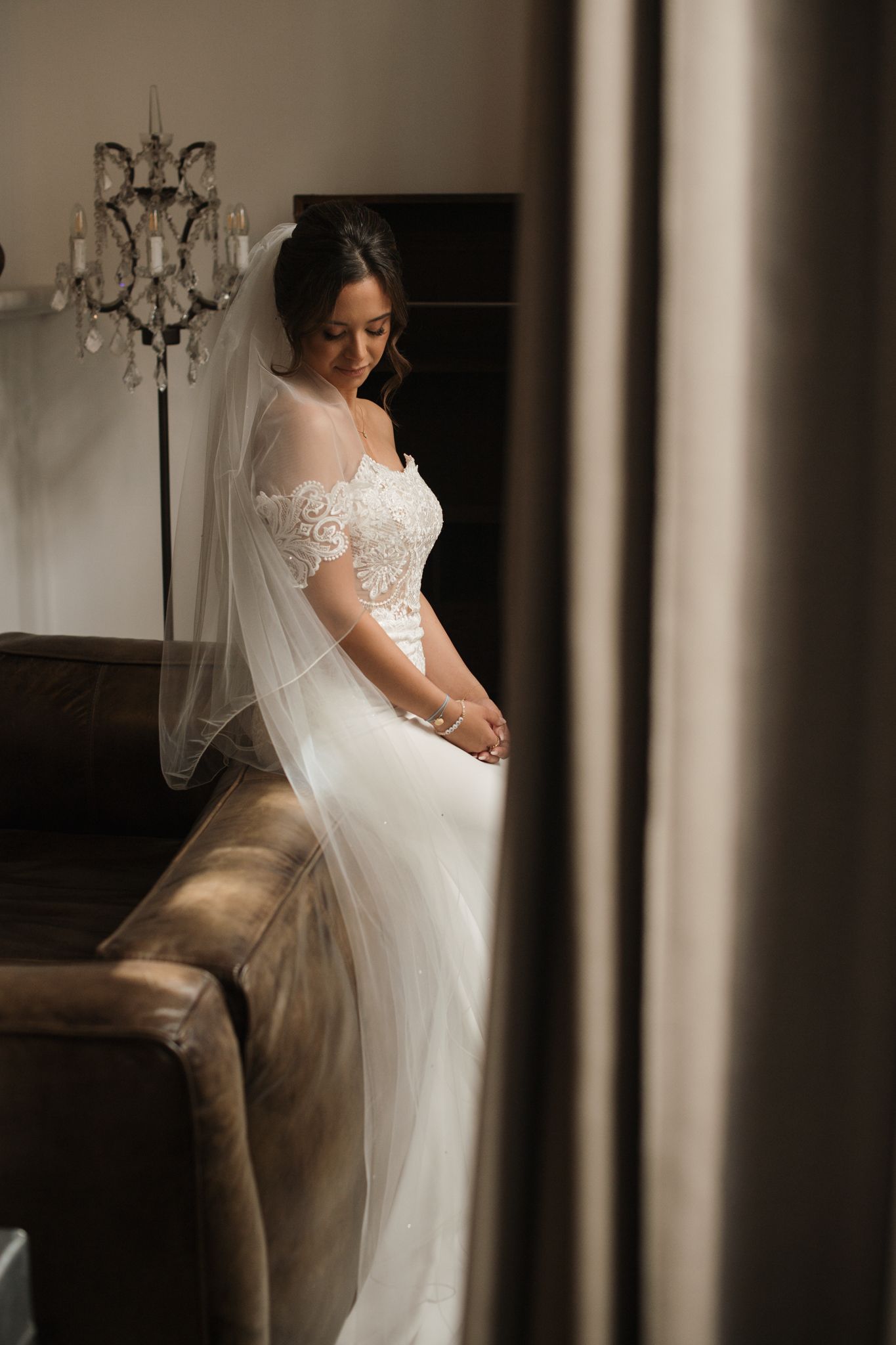 Beautiful bride in lace wedding gown and veil sitting on a leather sofa in a dimly lit room.