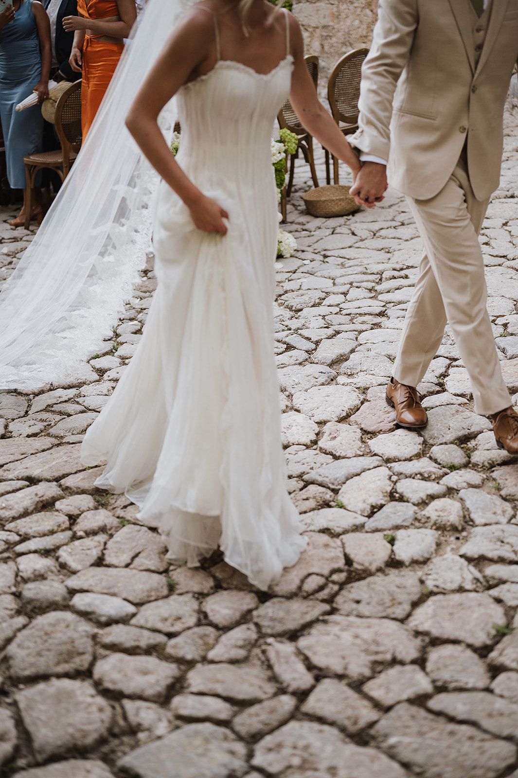 Bride in strapless white gown and groom in beige suit walk hand in hand on cobblestone path.